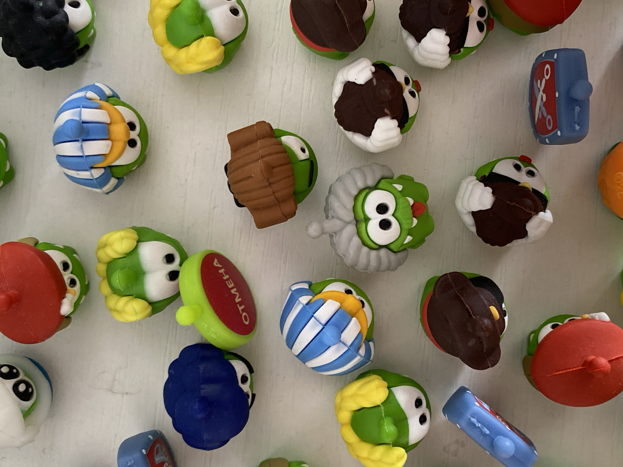 The image is a top-down, close-up shot of a collection of small, brightly colored toy figurines scattered on a white, textured surface. The figurines appear to be character toys, many of which are green with large eyes and some have accessories like hats or stripes. Some are wearing red hats, some are wearing brown hats, and one is wearing a blue, striped outfit. Another figurine has a red cap with white lettering that reads "ОТМЕНА". There is also a blue toy that resembles a digital alarm clock. The arrangement is random, with some figurines overlapping. The lighting is even, casting minimal shadows. There is no indication of any specific location in Ostrovki, Russia, or any human presence in the image.