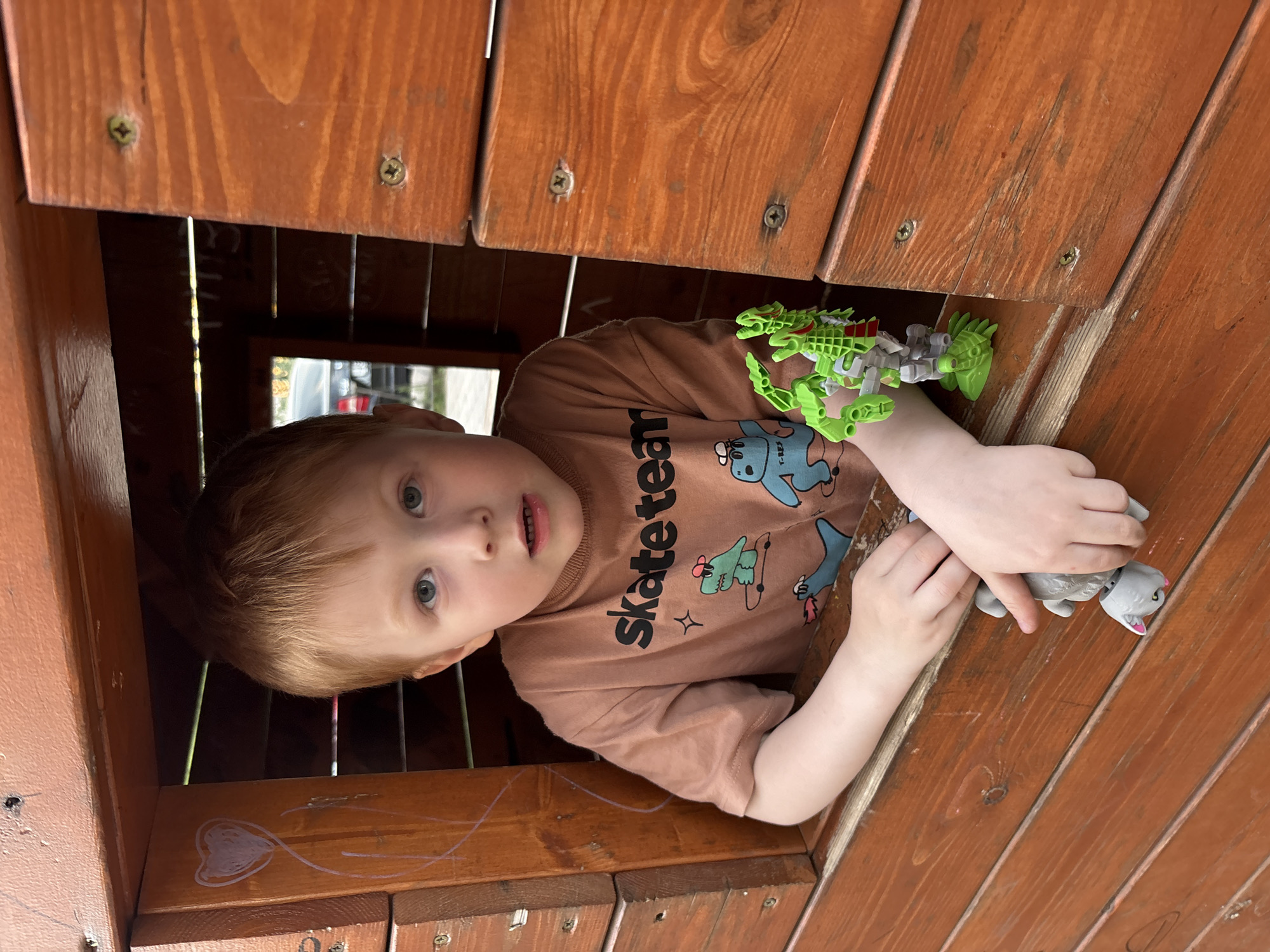 A young, fair-skinned child with reddish hair and green eyes is peering out of a wooden structure. The child wears a brown t-shirt with cartoon images and the word "Skate Team" printed on it. They are holding a green articulated toy in one hand and a small, gray toy animal in the other. The wooden structure appears to be part of a playhouse or fort, with visible wood grain and screws. A simple drawing of a heart and line is drawn on the wood below him. The scene has natural daylight. No information about Izhevsk, Russia is visible.