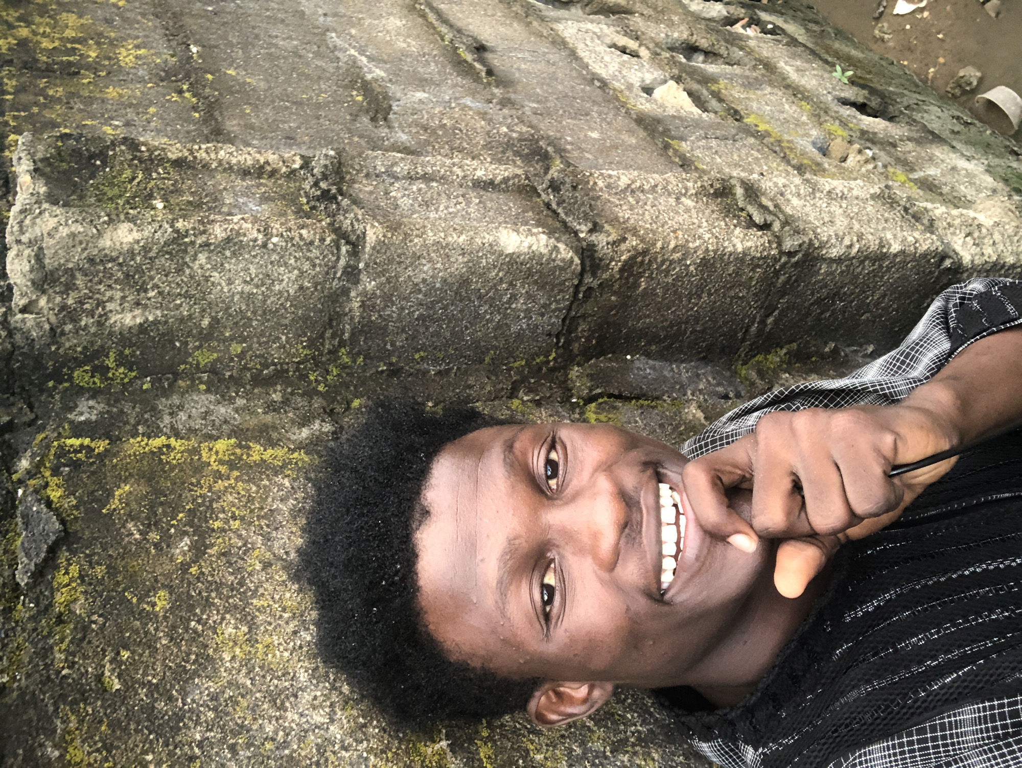 A young man of African descent, with dark, textured hair and a brown complexion, is positioned in the foreground, leaning against a rough, moss-covered concrete wall. He is looking directly at the camera with a wide, toothy smile. His right hand is raised, with his fingers gently touching his chin, and he appears to be in deep thought or contemplation. He is wearing a black and white checkered shirt with vertical striped accents on the collar and sleeves. The background is dominated by the weathered concrete wall, which shows signs of age and exposure to the elements, with patches of green moss growing on its surface. The lighting suggests an outdoor setting, possibly during the day. The scene appears to be located in Ado, Nigeria, based on the context provided.