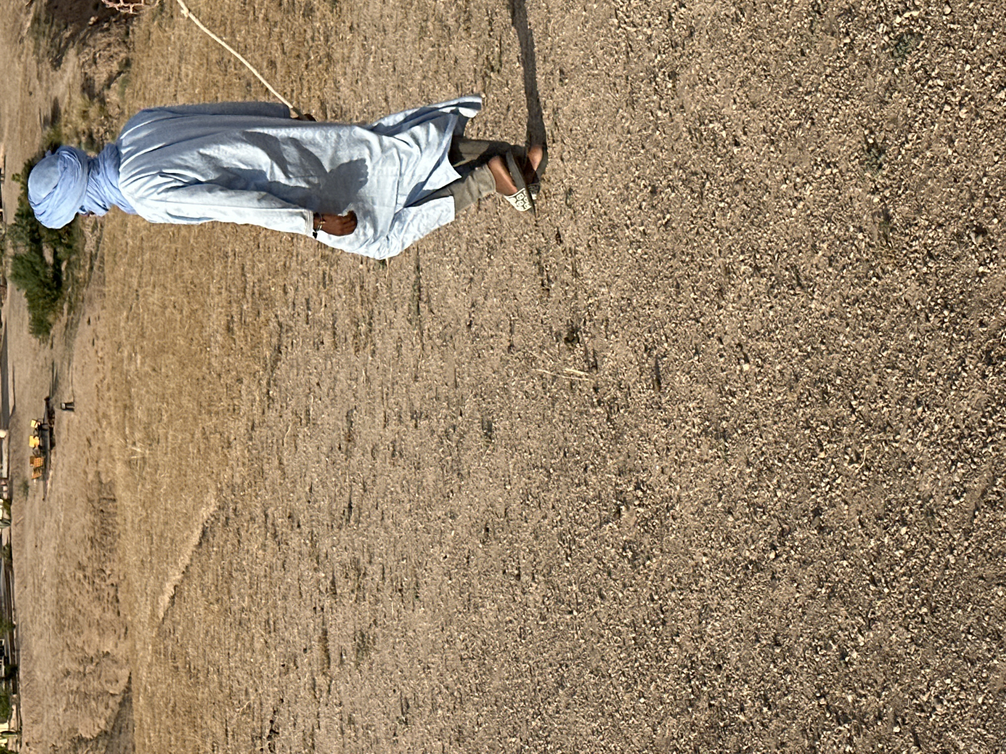 A man wearing a blue turban and a light blue garment stands in a dry, arid environment. The background suggests a location near the city of Sidi Badhaj, Morocco, based on the arid landscape. The ground is a mix of gravel and dry earth. A rope is visible. The person is wearing sandals or slip-on shoes.