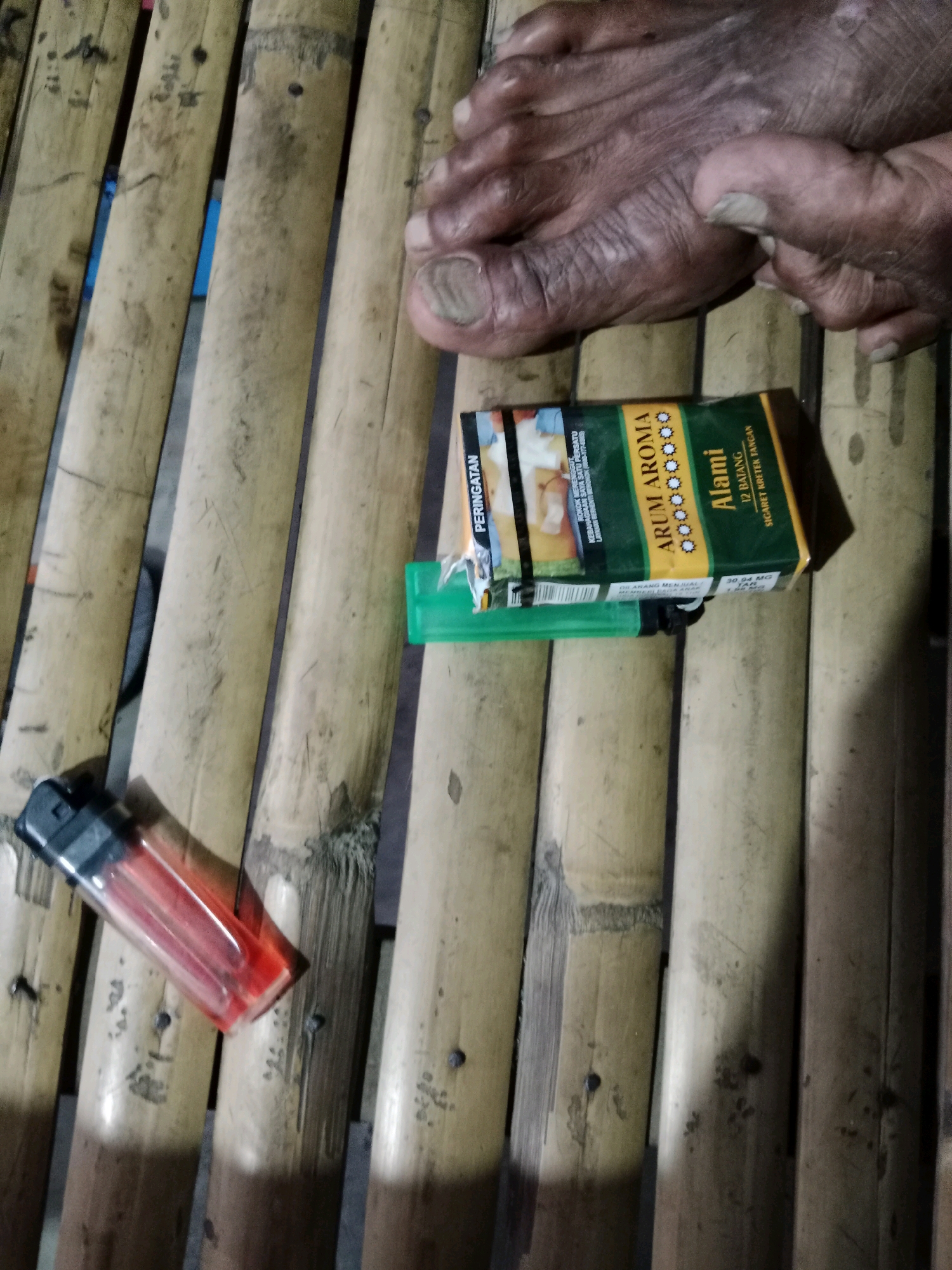 In Ringinrejo, Indonesia, a close-up shot captures a person's foot resting on a bamboo mat. A pack of "ARUM AROMA Alami" cigarettes, accompanied by a green lighter, lies on the mat near the foot. The scene is illuminated by artificial light, casting shadows on the bamboo surface. The perspective is from above, looking down at the objects and the foot.