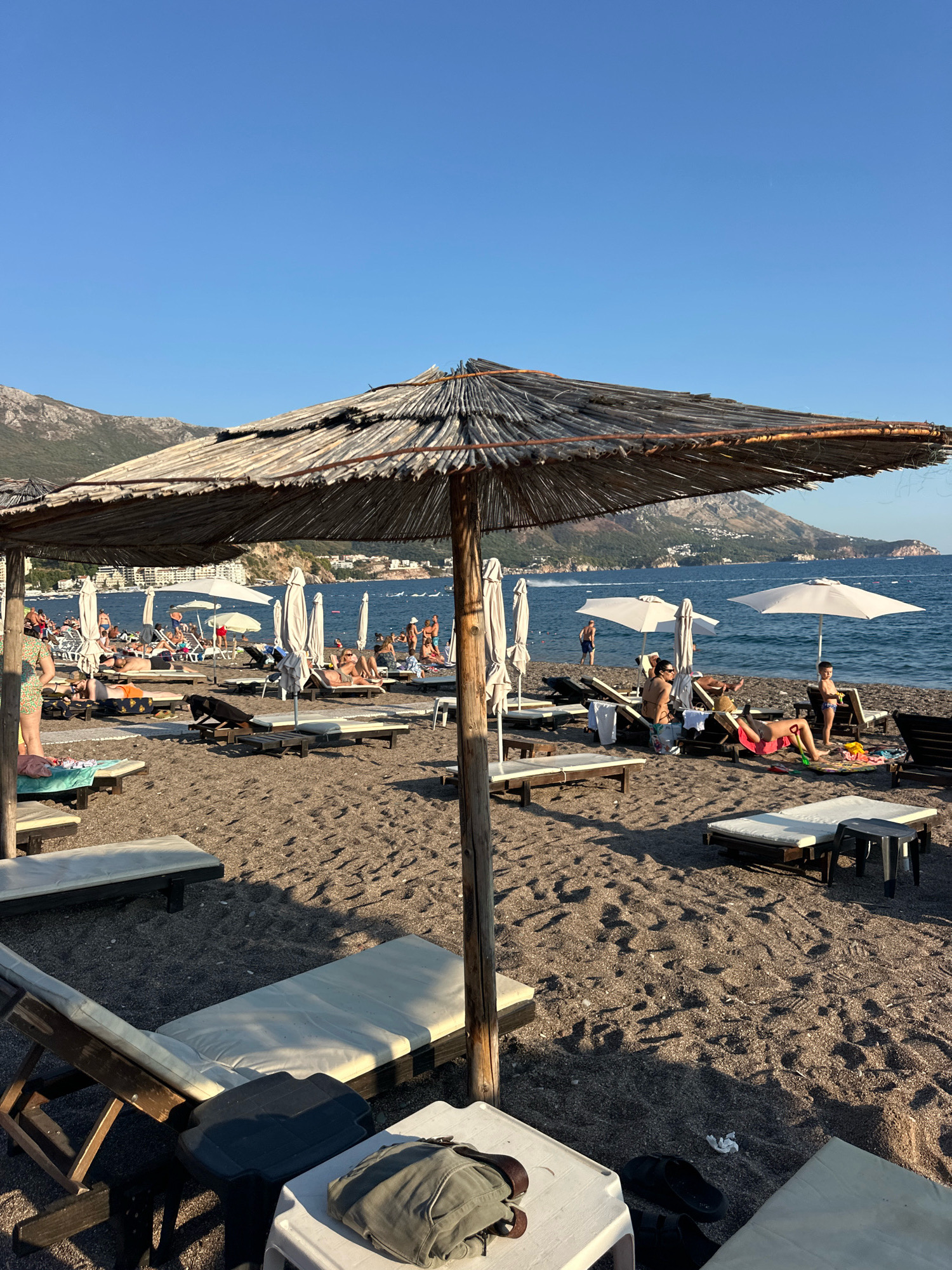 The image captures a busy beach scene in Boreti, Montenegro, under a clear blue sky. In the foreground, a weathered thatched umbrella casts a shadow over a sandy beach dotted with numerous lounge chairs, some occupied by sunbathers. Several folded white beach umbrellas are also visible. The dark sand of the beach transitions to the calm, blue sea in the background. In the distance, a coastal town with buildings is nestled against a backdrop of green, tree-covered mountains. Several people are scattered across the beach, some lounging on chairs, others standing and walking. Two white umbrellas are open near the water's edge, providing shade for those beneath them. The overall scene depicts a lively and typical summer day at a popular seaside resort.
