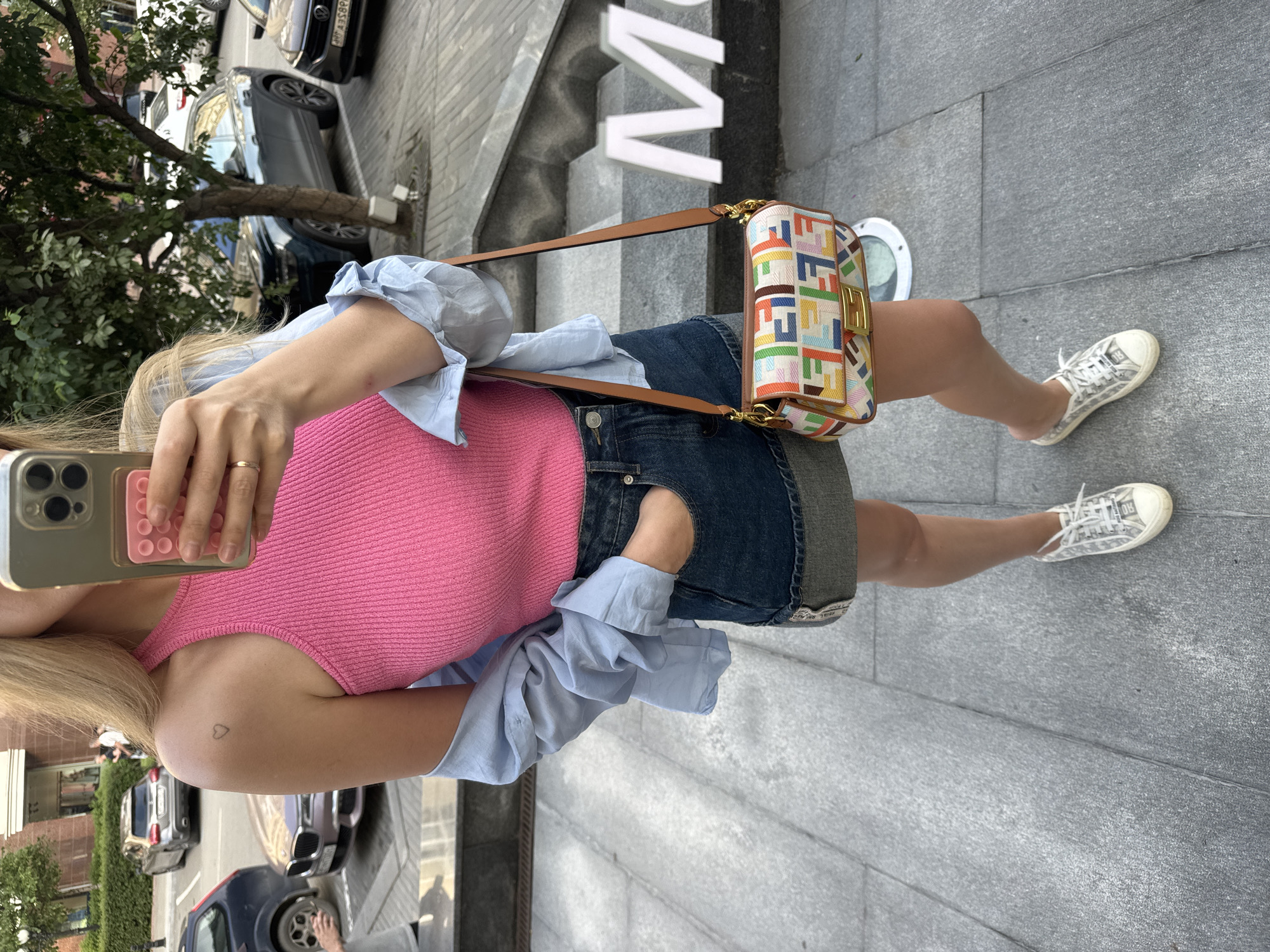 A person is captured from the waist down, holding a phone in their right hand to take a selfie. They are wearing a bright pink tank top, a light blue collared shirt tied loosely around their shoulders, and a denim skirt. White sneakers with a subtle pattern complete the outfit. A small, colorful Fendi bag with a brown strap is slung across their body. The person has a small heart tattoo on their left shoulder.
The background suggests an urban setting, with a glimpse of cars parked along a street and a building with large white letters forming the word "M". The ground is paved with gray stone tiles. The lighting indicates it is daytime, likely with clear weather. The overall mood appears casual and relaxed, with the person seemingly enjoying an outdoor stroll. The visible text includes the brand name "FENDI" on the bag and "M" on the building.