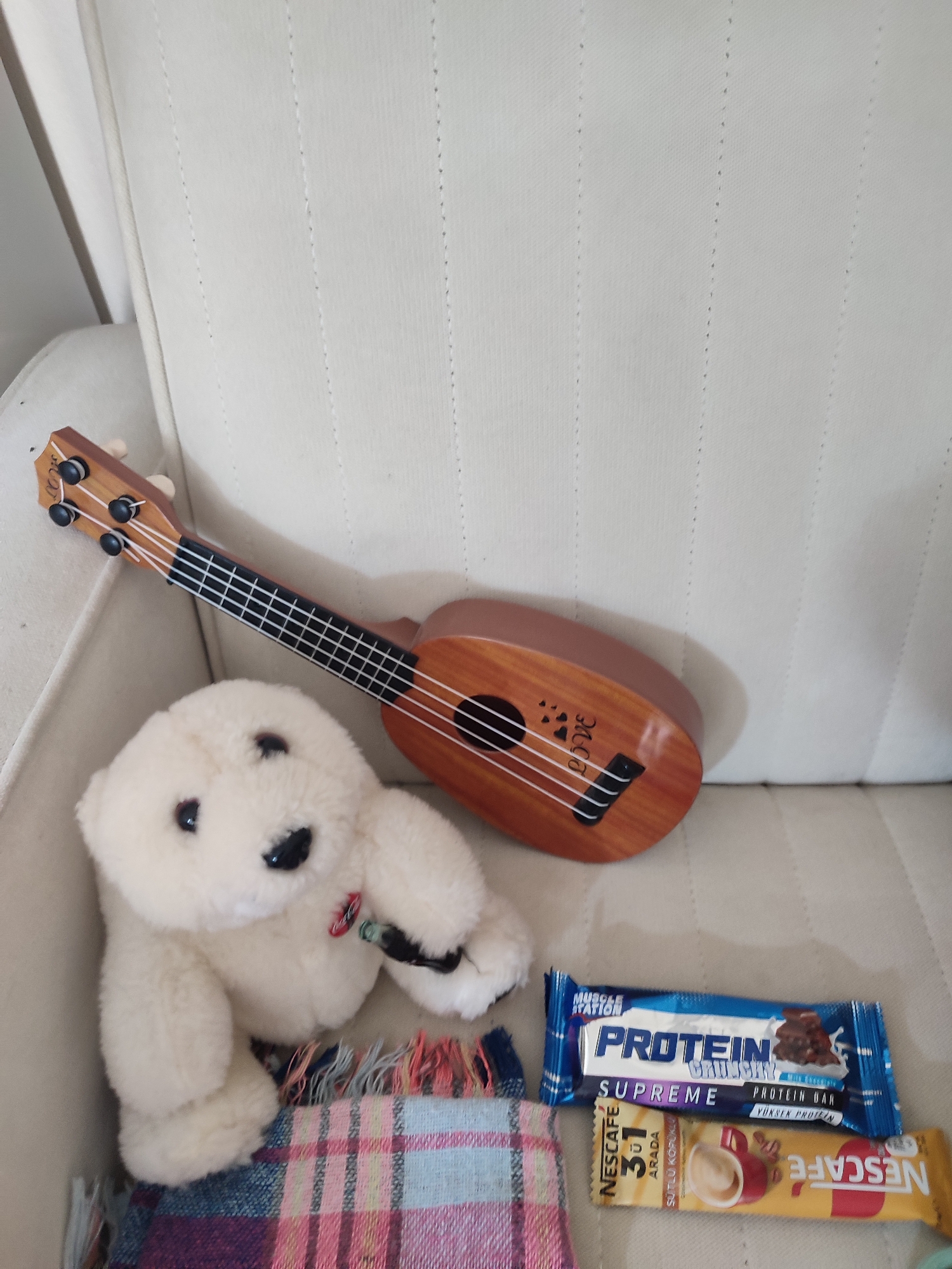 A still life image displays a plush white teddy bear positioned on a textured cream-colored sofa. To the right of the teddy bear lies a miniature wooden ukulele. Below the teddy bear and ukulele are two snack bars: one with blue packaging labeled "PROTEIN SUPREME" and another yellow package with the "NESCAFE" logo. A plaid blanket is partially visible beneath the teddy bear and snack bars. The scene is devoid of human presence and action. The image does not provide any information indicating its location in Üsküdar, Turkey.
