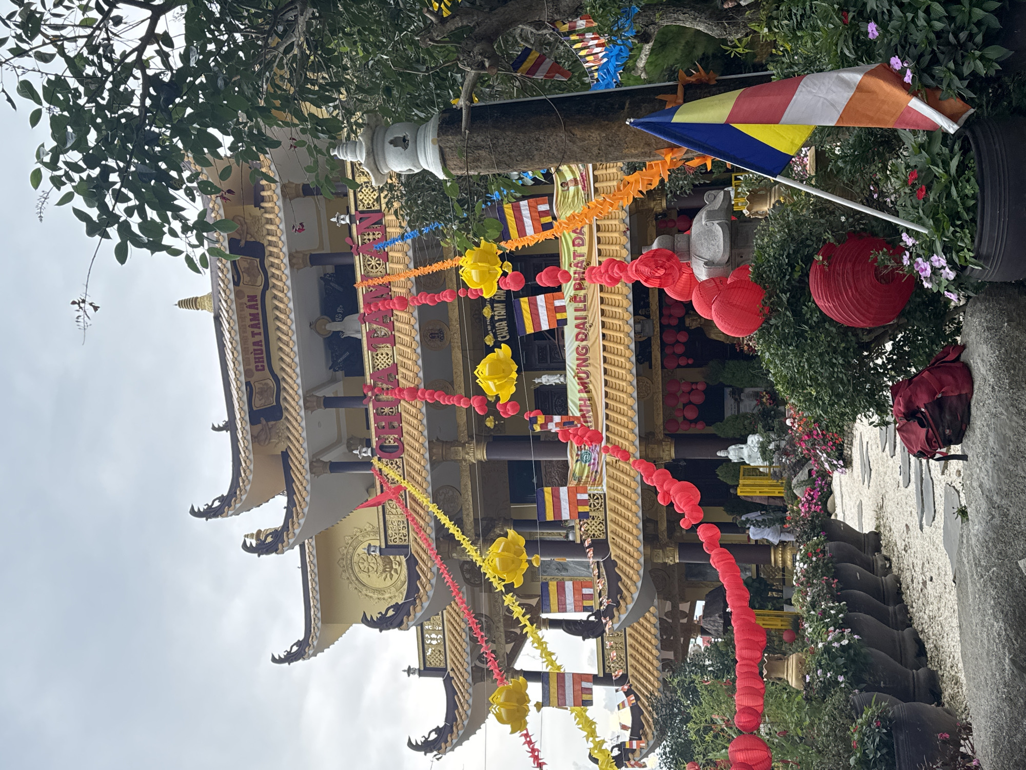 The image shows the exterior of Chua Tam An, a Buddhist temple located in Da Lat, Vietnam. The temple is a multi-tiered, ornate structure with yellow roofs and intricate architectural details, adorned with colorful paper lanterns and Buddhist flags. In the foreground, a pathway made of stones and gravel leads towards the temple entrance. Potted plants and flowers line the path, adding to the serene atmosphere. A red backpack lies on the ground near the pathway. Several people are visible in the background, some tending to the grounds and others entering the temple. The sky is overcast, suggesting a cloudy day.