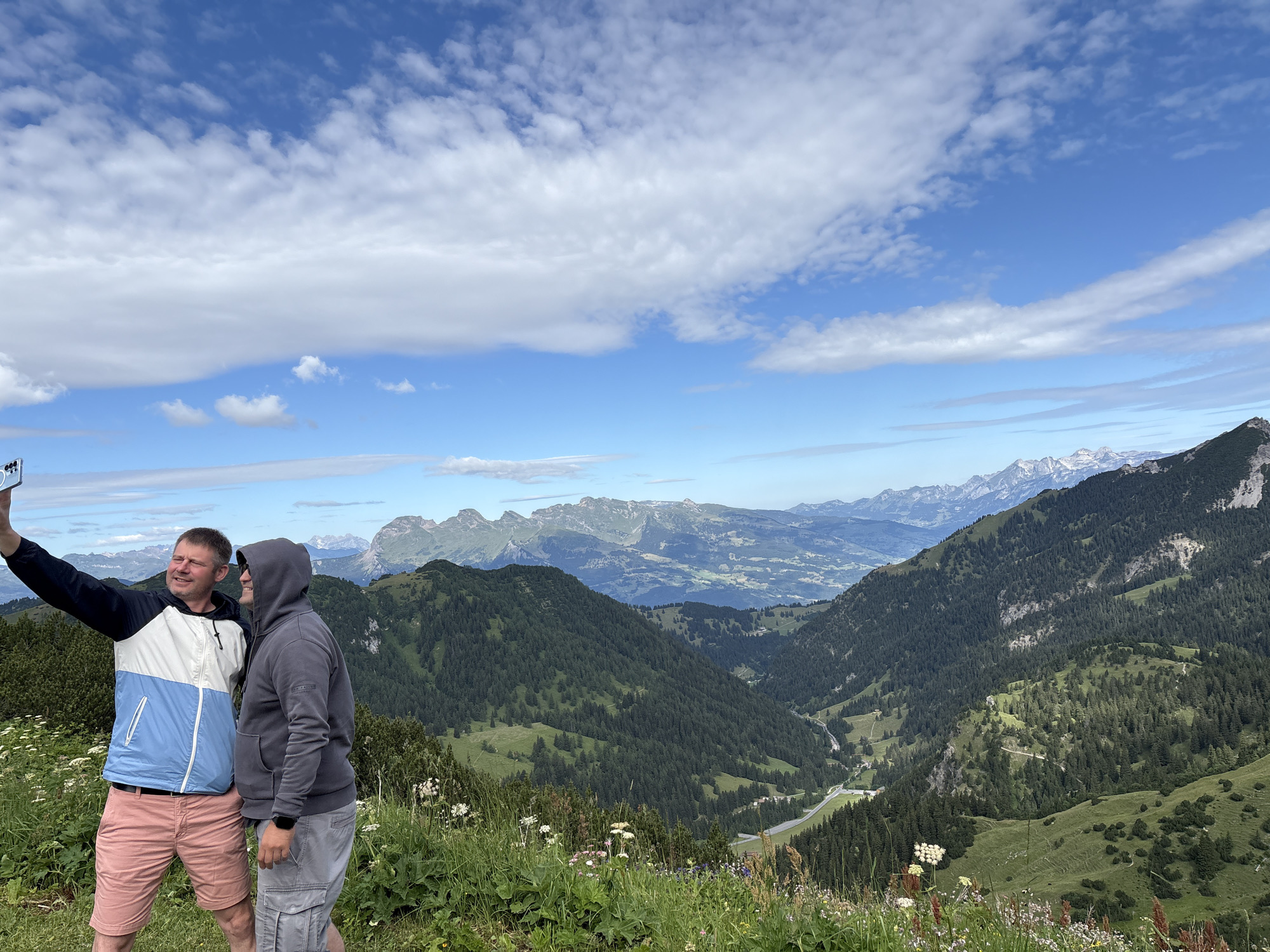 Two men are taking a selfie on a grassy hillside with mountains and a valley in the background. The man on the left is wearing a blue and white jacket and pink shorts, and is holding a phone in his left hand. The man on the right is wearing a gray hooded sweatshirt and gray cargo shorts. The man on the right is leaning into the man on the left, and both are smiling. The hillside is covered in green grass and wildflowers. In the background, there are rolling green hills covered in trees, and further in the distance, there are snow-capped mountains. The sky is blue with scattered white clouds. This appears to be a scenic overlook in Malbun, Liechtenstein.