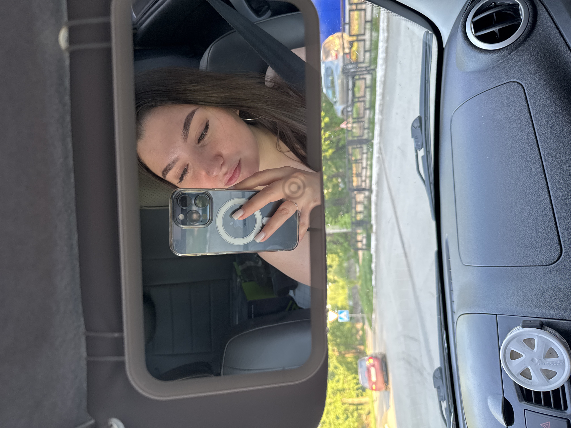 A woman is taking a selfie in the rearview mirror of a car. She's wearing a seatbelt. The image reflected in the mirror shows a street scene with trees and a red car. Based on the reflection, the location is Perm, Russia. The interior of the car is visible, including the dashboard and air vent. The phone in her hand has a MagSafe charger attached.