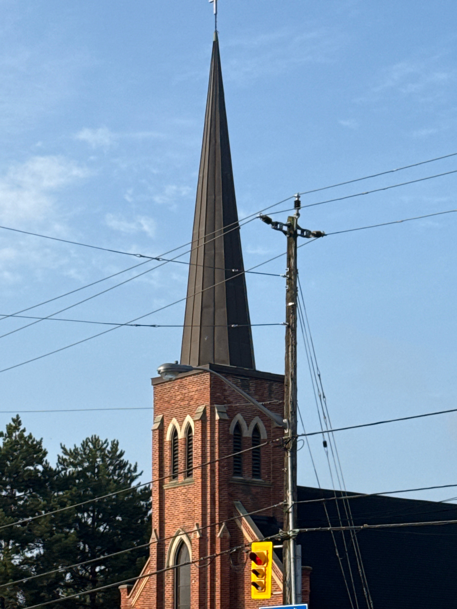 This image, taken from a low angle, shows the top portion of a red brick church with a tall, dark spire topped with a white cross. The spire is prominent and reaches high into a clear blue sky with sparse, wispy clouds. Multiple power lines crisscross the frame, obscuring parts of the church and the sky. To the right of the church spire, a weathered wooden utility pole stands tall, also strung with numerous wires. Below the pole and partially in front of the church's lower section, a yellow traffic signal with red and amber lights is visible. Evergreen trees can be seen to the left of the church, and a dark, sloping roof of an adjacent building is visible in the lower right quadrant. The overall impression is an urban landscape with a religious building set against a backdrop of infrastructure and natural elements.