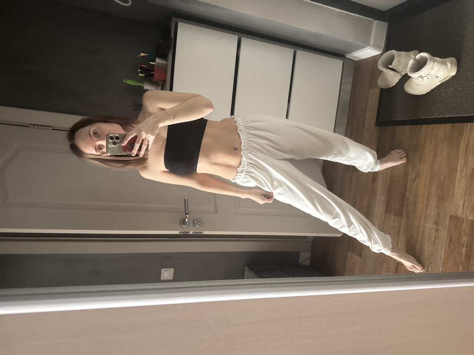 A woman is taking a selfie in a bathroom mirror. She is wearing a black strapless top and white drawstring pants. Her brown hair is shoulder-length. In the background are white cabinets, a black wall with some containers, and white sneakers on a mat on the wood floor.