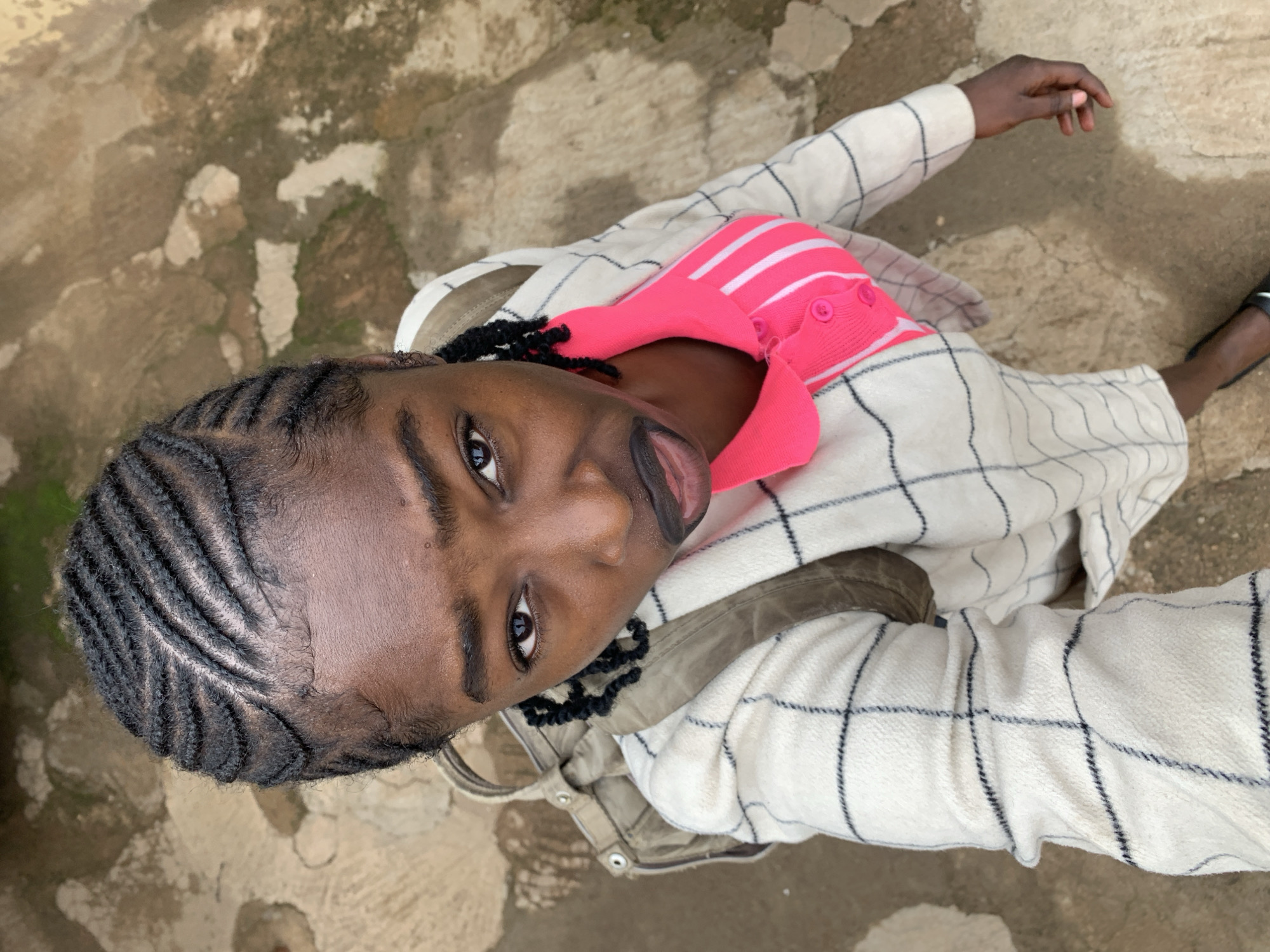A young Black female stands outdoors in Rafin Par, Nigeria, captured in a high-angle shot looking directly up at the camera. Her hair is styled in tight cornrow braids, meticulously arranged in parallel lines that curve across her scalp from front to back. She wears a vivid pink collared top featuring white horizontal stripes on the upper chest and three pink buttons down the placket. This is layered under an off-white, long-sleeved coat with a dark plaid grid pattern. A light brown canvas backpack is visible over her left shoulder. Her right hand is partially visible, resting at her side, and a portion of a dark flat shoe is seen on her right foot. The environment consists of an uneven, light brown-tan ground, appearing as cracked, worn concrete or hardened earth, interspersed with darker patches of dirt or moss.