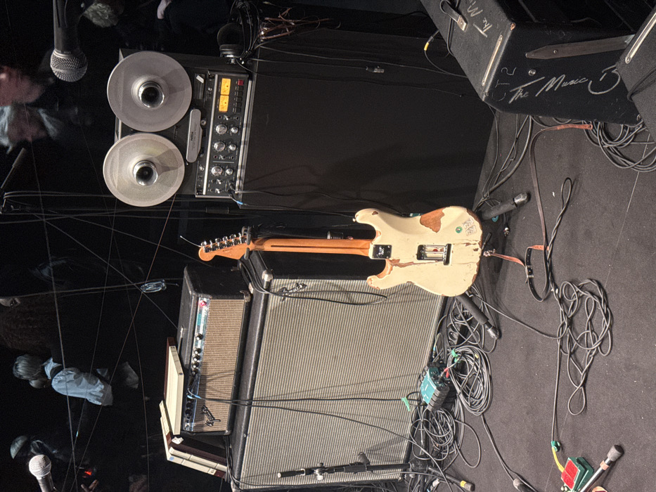 The scene depicts a detailed musical performance or recording setup in Paris, France. A cream-colored electric guitar, heavily worn and relic'd on its reverse, stands vertically on a floor stand. To the guitar's left, a large silver-grille Fender speaker cabinet is stacked with a smaller black Fender amplifier head, on which a light-colored rectangular object and several books rest. To the guitar's right, a TASCAM 388 reel-to-reel tape recorder with two active metallic reels sits atop a rack-mounted audio mixer/processor unit featuring multiple controls and illuminated VU meters. Black headphones hang from this unit. The floor is covered in dark carpeting and is extensively cluttered with numerous black audio cables, coiled and sprawling, along with several microphones on stands and a green/black audio interface. A brown leather guitar strap lies on the floor. In the lower right, a black equipment case or speaker cabinet with "The Music" and "CT2" written in white is partially visible. The background is dimly lit and out of focus, revealing indistinct figures of people, suggesting an audience or crew in an indoor venue. Thin cables and wires are strung across the dark background.