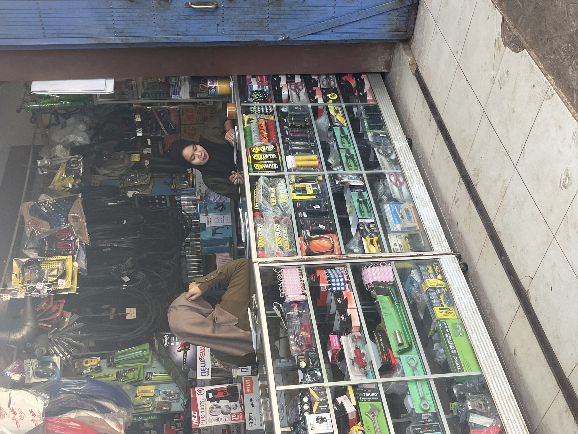 A two-person shop displays automotive parts and tools. A person wearing a hijab is standing behind a counter with their hand near their head, looking to the left. Another person in a hijab is seated behind the counter, looking down at documents and a keyboard. The shop is filled with shelves of neatly organized items, including boxes of tools, spare parts, and accessories. The store appears to be located in Karang Sari, Indonesia. The storefront has a blue metal door and the interior is lit by overhead lights. The floor is tiled with a dark metal strip running across it.