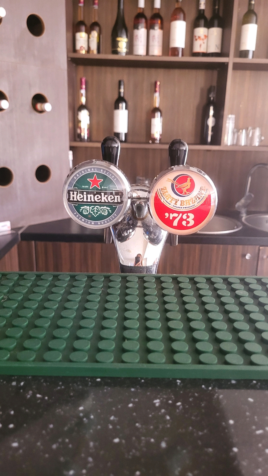 This is a close-up, eye-level, vertical shot of two beer taps on a bar. The tap on the left has a circular emblem with the Heineken logo, featuring a red star and the brand name. The tap on the right has a circular emblem with a red background, a golden pheasant graphic, the text "PLATTY" and "GOLDEN PHEASANT", and the number "73". Both taps are made of polished metal, and the handles appear to be black. They are positioned in front of a green, plastic mat with a textured, circular pattern, which is likely a drip tray. Behind the taps, shelves stocked with various bottles of alcohol are visible, suggesting the setting is a bar or pub. The background is blurred, but includes wooden shelving and some glassware. There is no indication of the specific location in Prešov, Slovakia.