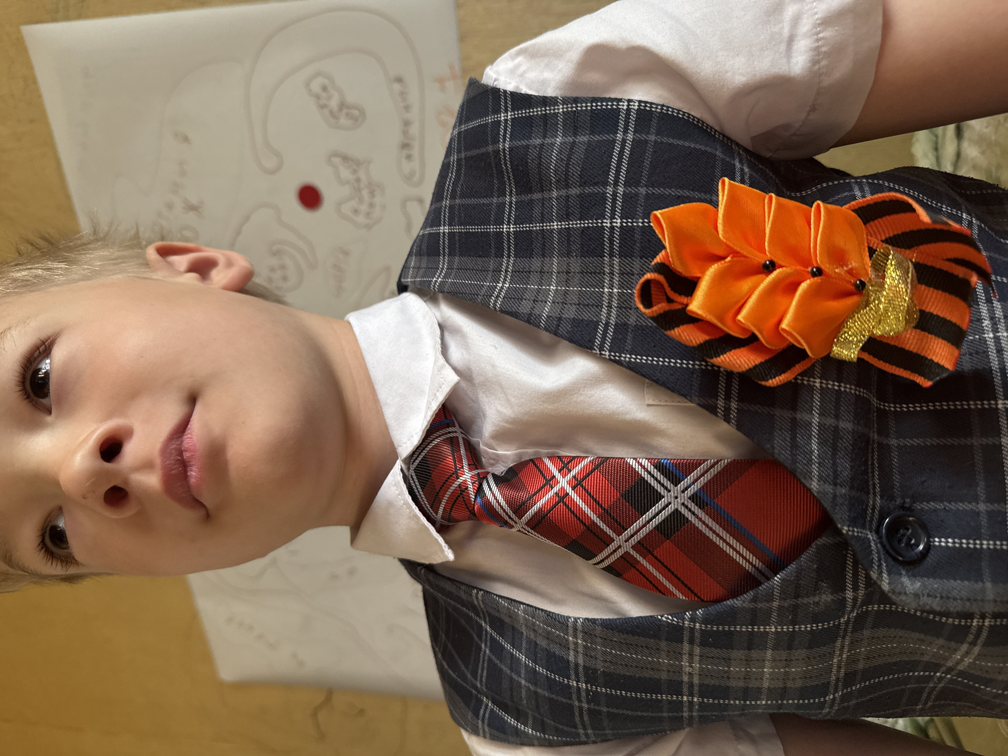 A child wearing a white collared shirt, a plaid tie, and a plaid vest is the subject of this close-up photograph. On the vest's lapel is a flower-shaped pin with orange petals and a St. George ribbon (black and orange stripes). A piece of paper with a drawing on it is in the background. The location is Dmitrov, Russia.