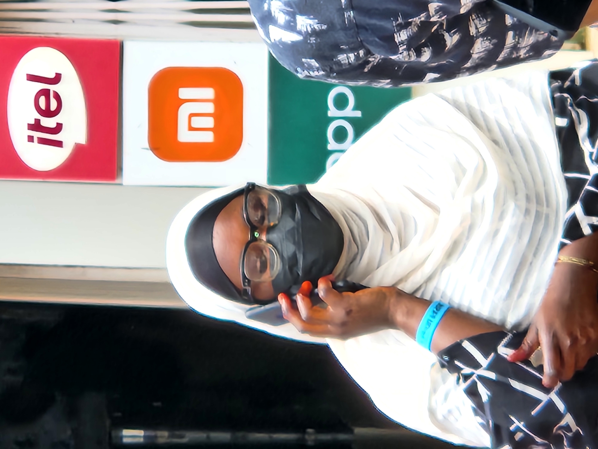 A medium shot captures a woman wearing a white hijab, a black face mask, and glasses, holding a smartphone to her left ear and speaking on it. She is adorned with a blue wristband on her left arm and a gold bracelet on her right wrist. She is dressed in a black and white patterned top. To the right, another person, partially visible, wears a black garment with a white abstract pattern. The background features signage with the "itel" logo on a red background and the "MI" logo on an orange background, followed by a green sign with the partial word "app" visible in white text. The scene suggests a street-level view in a commercial area.