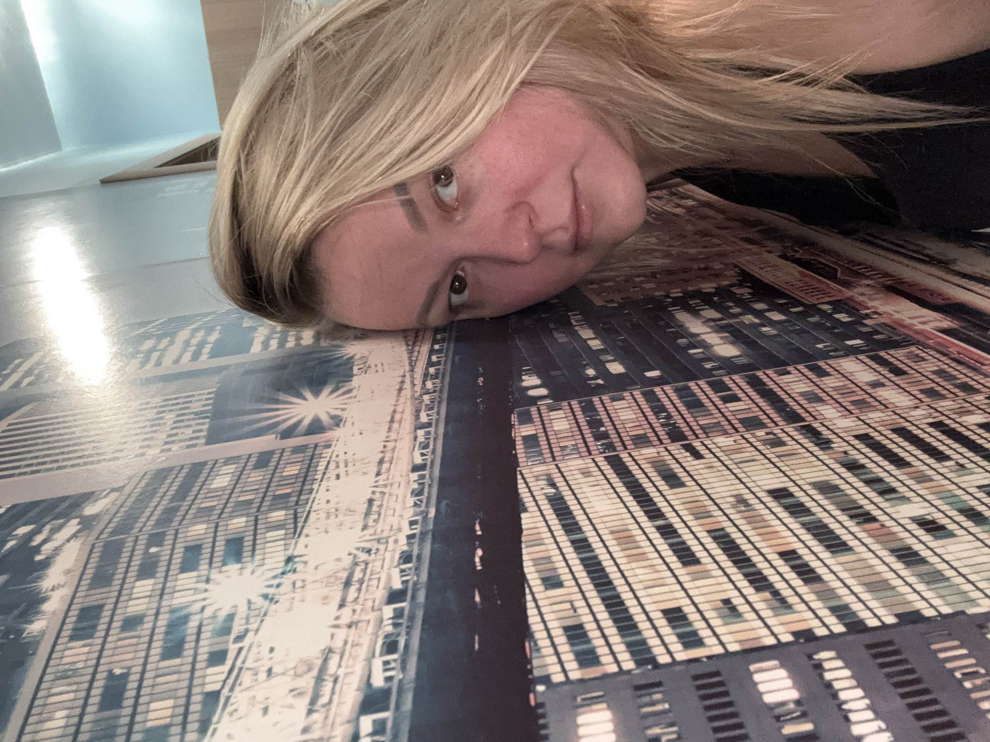 A blonde female individual, estimated to be in her 20s to 30s, is depicted in a close-up, head-and-shoulders portrait, leaning her head against a wall in what appears to be an indoor setting in the city of Русский Вожой, Russia. The wall is covered with a large mural or wallpaper that illustrates a nighttime cityscape, featuring numerous illuminated skyscrapers and an elevated structure resembling a bridge with various lights. The female subject's right cheek (viewer's left) is pressed against the mural. Her long, light-blonde hair is parted and flows around her shoulders. She is wearing a black sleeveless top. Her brown eyes are open and directed at the camera, maintaining a neutral expression. The mural displays a cool color palette with prominent artificial lights across the buildings and bridge, some exhibiting starburst effects. A segment of a light blue wall or ceiling is visible in the upper right background, beyond the mural. The scene indicates a posed photograph.