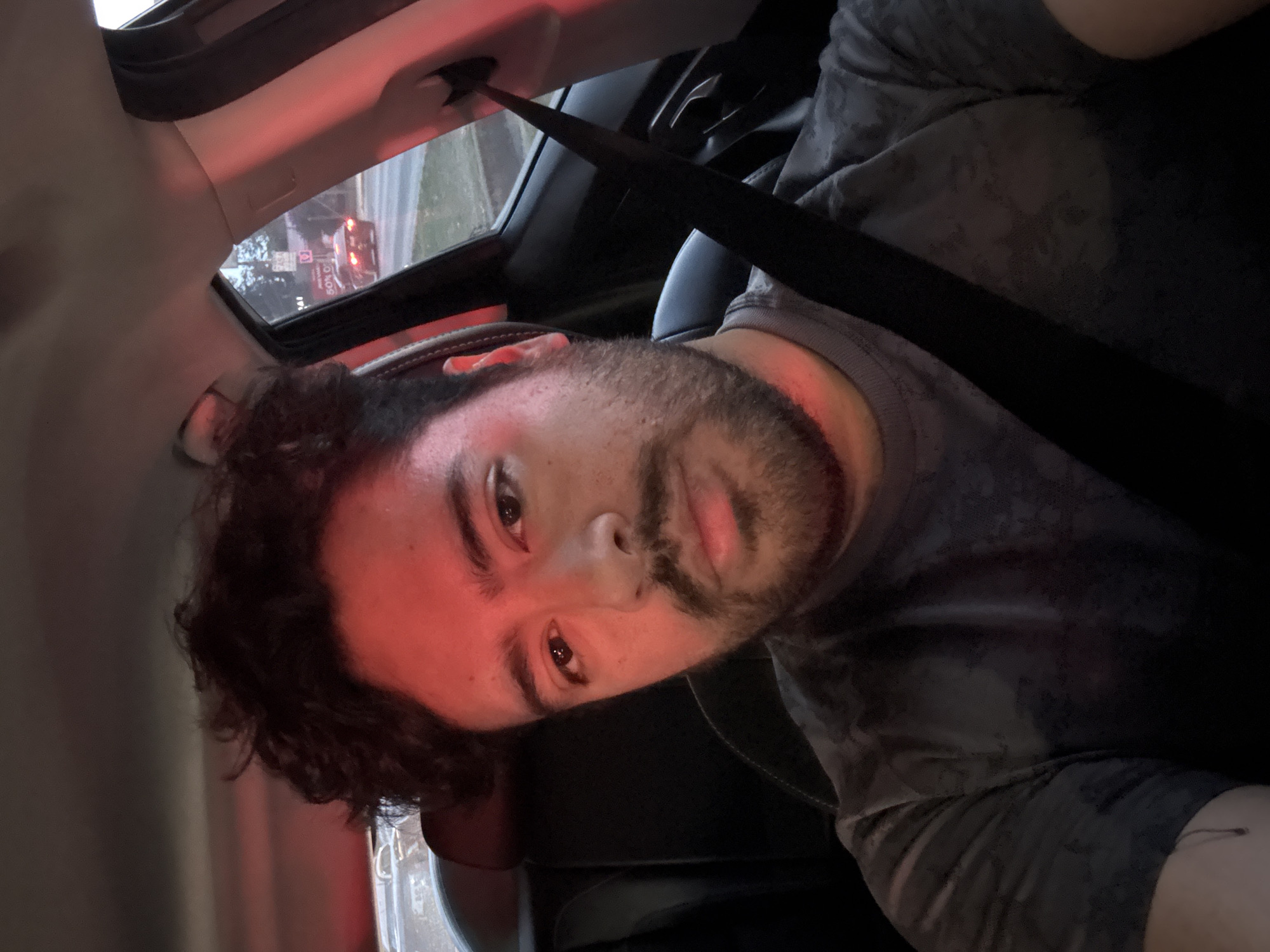 A selfie-style portrait captures a male individual inside a car, likely at dusk or night, in Puebla, Mexico. The subject, a man with dark curly hair and a beard, is looking directly at the camera with a neutral expression. He is wearing a dark, possibly patterned, t-shirt and has a seatbelt fastened across his chest.
A prominent red glow illuminates parts of his face, the car's interior, and is cast from outside the vehicle, suggesting he is stopped at a red traffic light. Through the sunroof, a dim sky is visible, along with distant streetlights or vehicle headlights, confirming the evening or night setting. Also visible through the sunroof is a commercial sign with the text "50%" clearly readable, indicating a discount or advertisement. The car's dark interior, including the seat and roof lining, provides a contrasting backdrop to the red light. The overall mood is calm, captured during a momentary stop in transit.