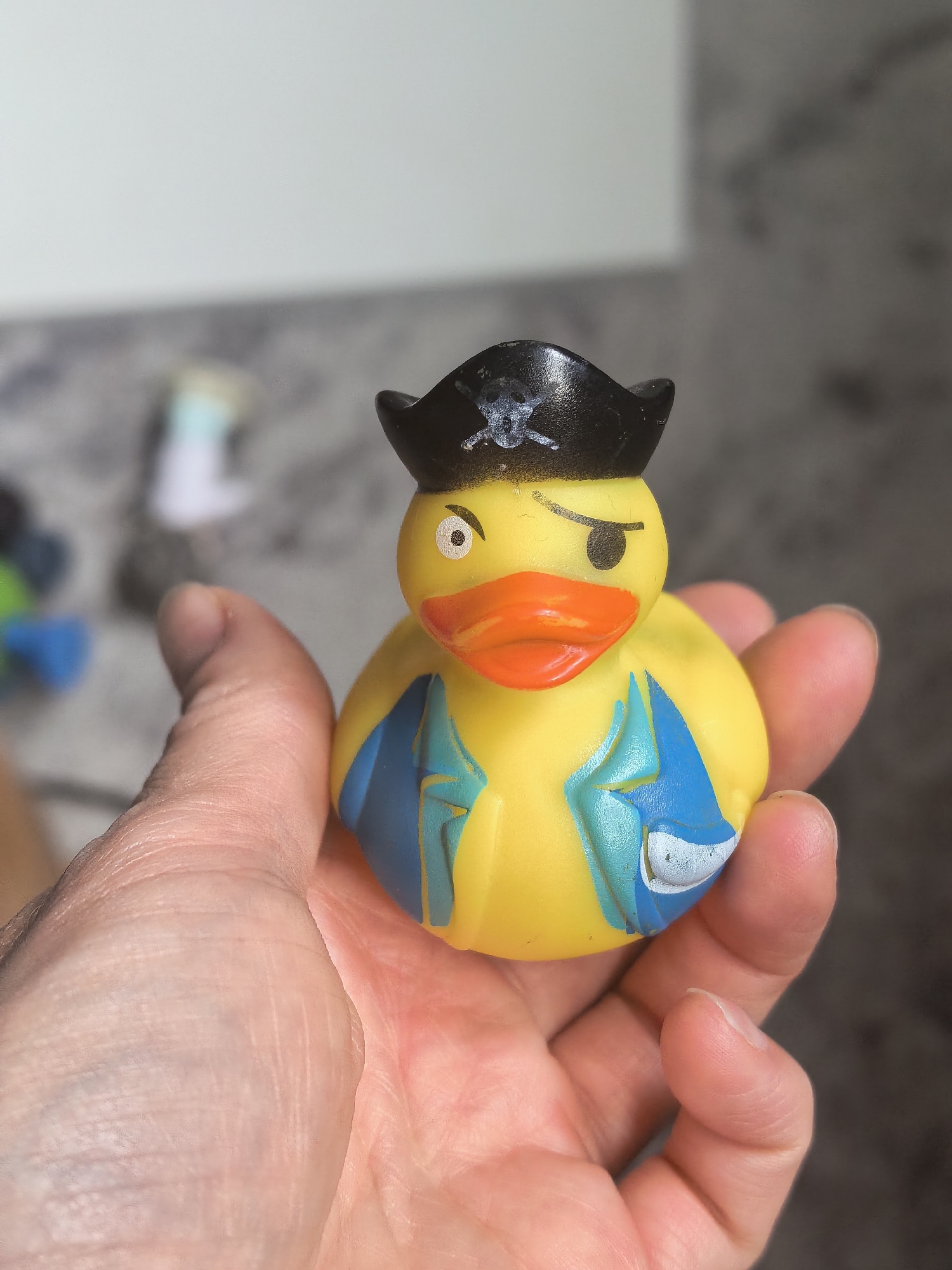 A close-up shot shows a yellow rubber duck toy, dressed as a pirate with a black hat and an eye patch, being held in a person's left hand. The background is blurred, with a hint of a bathroom or laundry room setting, including what appears to be a grey countertop and possibly a washing machine. There is no indication of the city of Sterlitamak, Russia, in the image.
