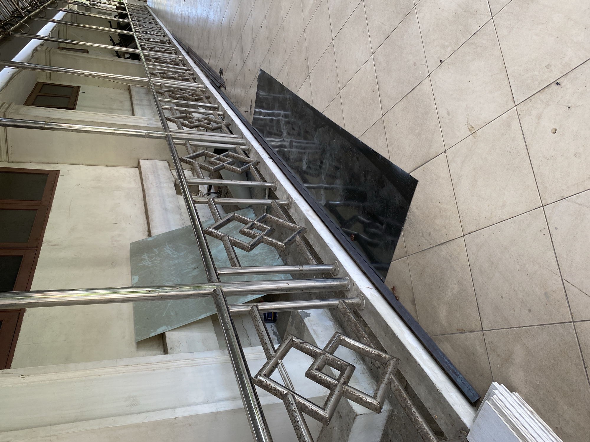 The image depicts an outdoor covered walkway or veranda. A metal railing with a geometric pattern runs along the length of the walkway, separating it from a lower area that appears to be a set of stairs. The wall behind the railing is constructed of light-colored material, possibly concrete, with dark-framed windows visible at intervals. The floor of the walkway is tiled with light-colored square tiles. A long, rectangular piece of black material, possibly a mat or a smooth surface, is placed on the tiled floor adjacent to the railing. In the background, obscured by the railing, are glimpses of what appear to be motorcycles parked. To the left of the walkway, near the stairs, are several stacked rectangular objects, likely tiles or building materials, and a large, light-colored board leaning against the wall. The overall scene suggests an unfinished or under-construction area within a building in Banda Aceh, Indonesia.