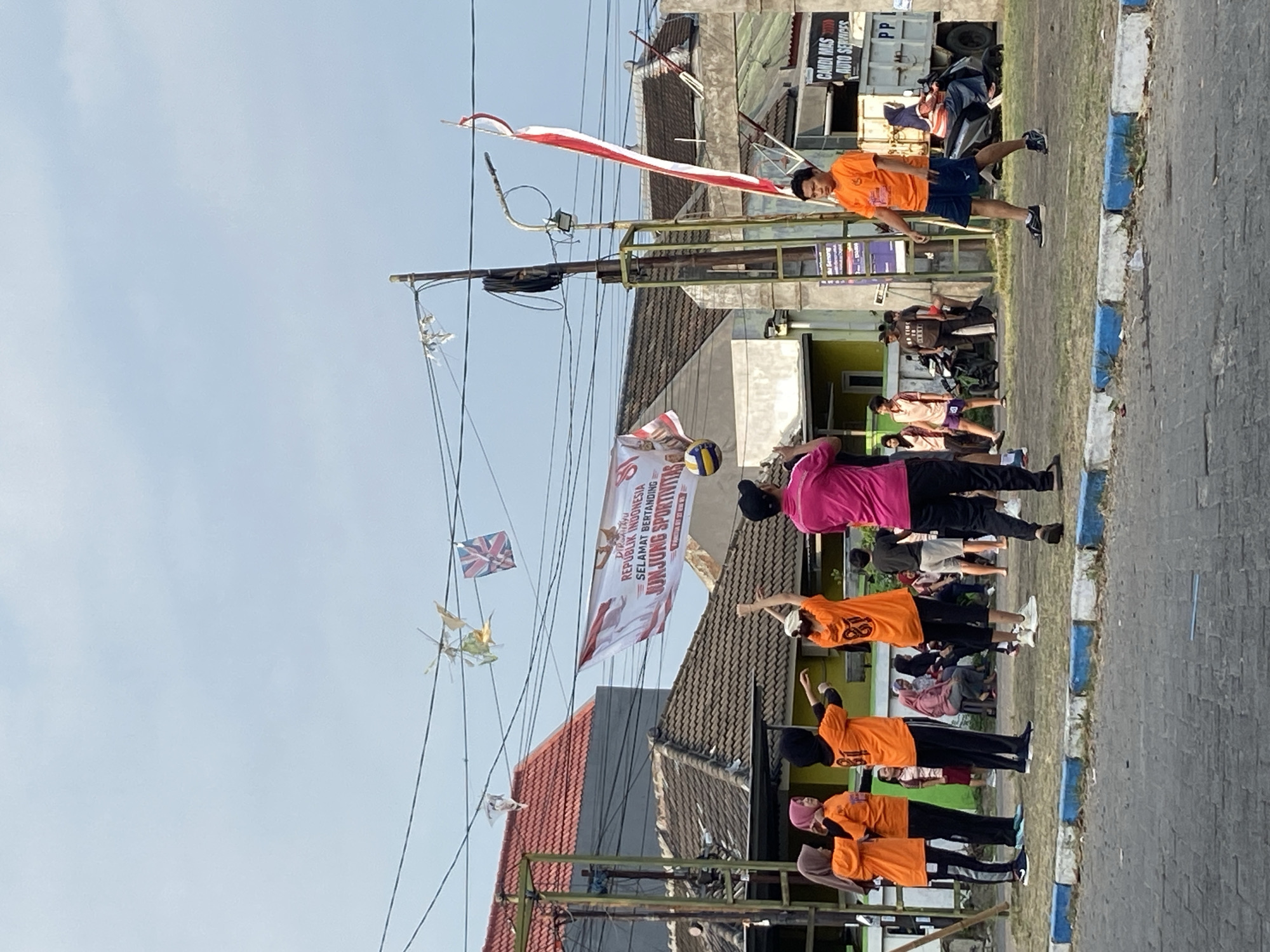 In the city of Gelam, Indonesia, a group of people are engaged in a volleyball game outdoors. The scene is characterized by a bright, overcast sky and a mix of residential and commercial buildings. Several individuals, predominantly wearing orange t-shirts with the number "81" on them, are actively participating in the game. One person in a pink shirt appears to be coaching or directing. The volleyball itself is visible mid-air, suggesting an ongoing rally. A banner with Indonesian text and graphics hangs across the scene, potentially indicating a local event or competition. Decorative flags, including what appears to be a Union Jack, are strung overhead, adding to the festive atmosphere. In the background, a white truck with "CANDI MAS AUTO SERVICES" written on it is parked, and motorcycles are visible, hinting at the mode of transportation common in the area. The ground is paved with asphalt and bordered by a curb with blue and white markings.