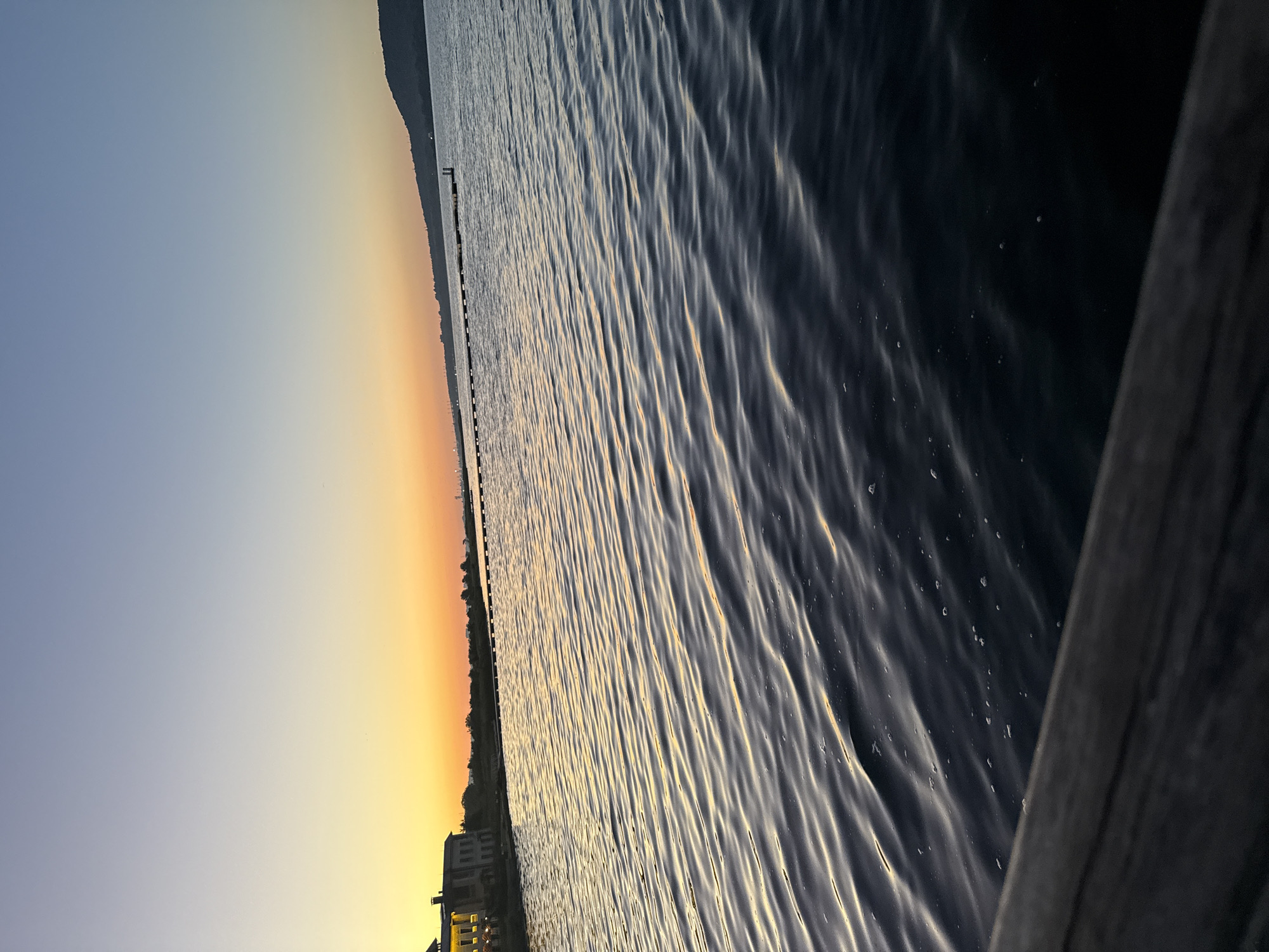 The image depicts a serene body of water at dusk, with a clear sky transitioning from a pale blue to a warm orange hue near the horizon. In the distance, a line of trees and structures is visible along the shoreline, and a long pier or breakwater extends into the water. A faint outline of a hill or mountain is present on the right side of the horizon. The foreground is dominated by the dark, rippling surface of the water, with reflections of the sunset catching the crests of the waves. A wooden railing or edge of a dock is visible in the lower left corner, partially obscuring the water. The overall atmosphere is peaceful and tranquil, suggestive of a quiet evening by the water. The specific location of Казашко, Bulgaria, is not visually identifiable from the image itself.