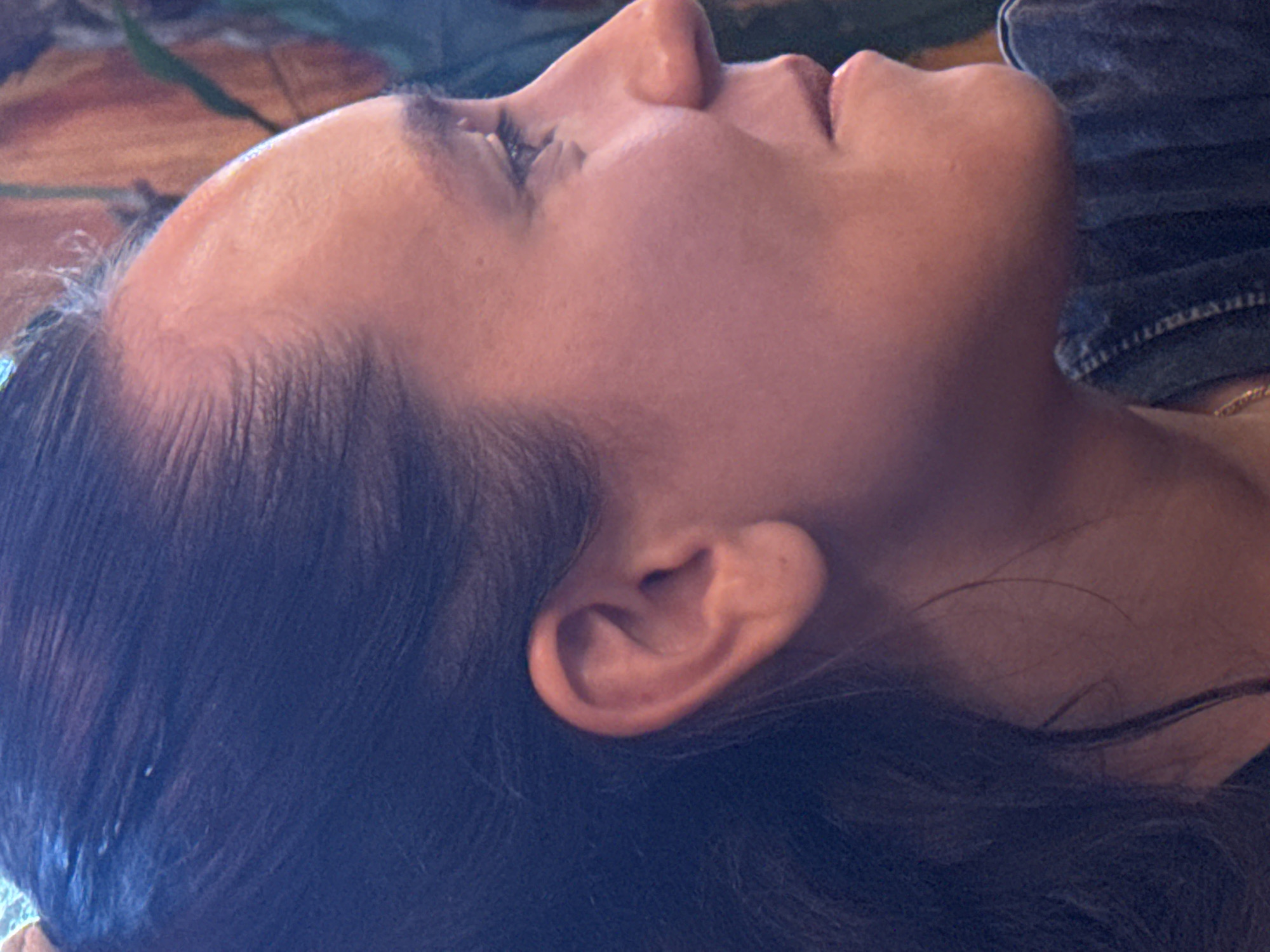 A close-up, high-angle shot of a light-skinned woman's face and upper torso, appearing to be lying down. She is turned to her right, with her face angled upward. Her dark hair frames her face and neck. The top of her head is visible, showing a receding hairline. Her ear is prominent in the mid-ground. She has a closed eye, a prominent nose, and slightly parted lips. A gold chain is partially visible around her neck. The background is slightly blurred, showing a mix of dark blue and what appears to be a patterned surface. No indication of Mèo Vạc Commune, Vietnam, is present in the scene.