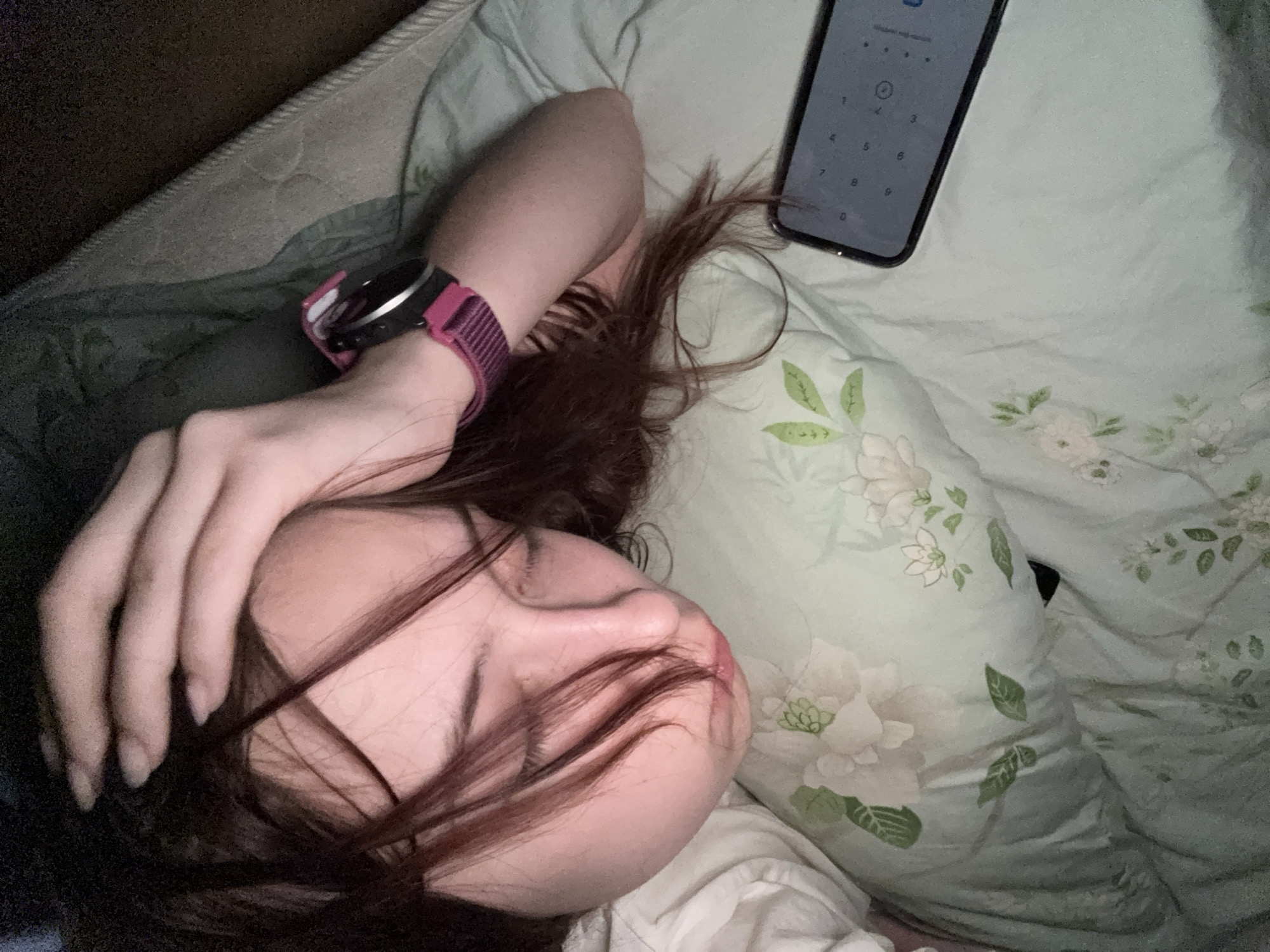 A person is lying on a bed, possibly sleeping. They are positioned on a floral patterned pillow and covered with a white sheet. The person has dark hair and is wearing a pink watch on their left wrist. Next to the person lies a smartphone displaying a number keypad. The environment appears to be a bedroom. There is no visible indication or mention in the image data that it is taken in Belaya Pashnya, Russia.