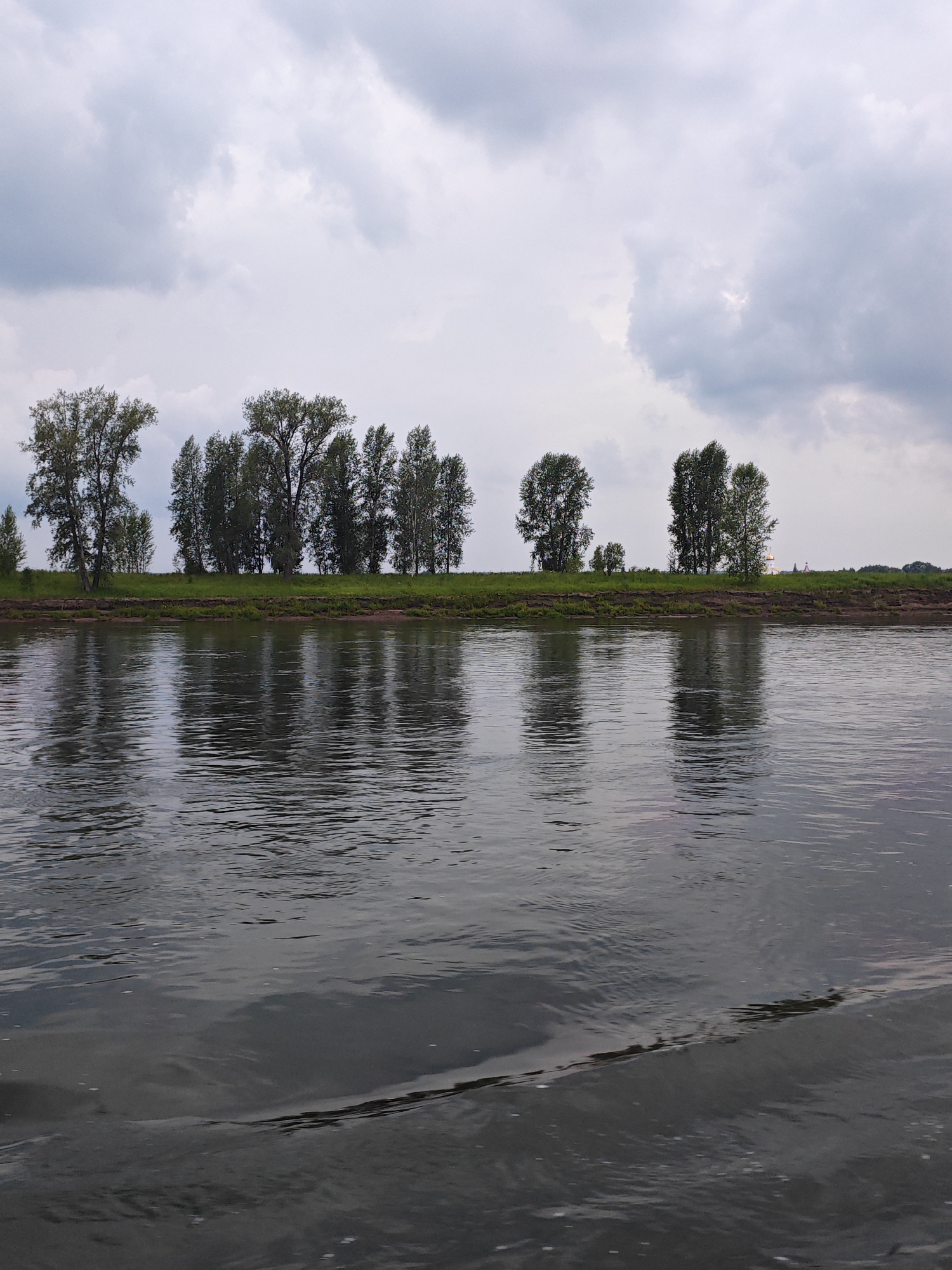 An overcast day is captured in this landscape image, showing a wide river in the foreground and a tree-lined bank in the distance. The river's surface is rippled, reflecting the muted grey sky and the dark green foliage of the trees. The trees are of various sizes and densities, forming a continuous line along the elevated bank. The bank itself appears to be covered in grass, with a darker strip of soil or sand visible at the water's edge. In the far distance, beyond the trees, hints of man-made structures are barely discernible, suggesting a settled area or perhaps a road. The overall impression is one of natural tranquility under a cloudy sky. The location in Kazachinskoe, Russia, situates this scene within a region known for its rivers and natural landscapes.