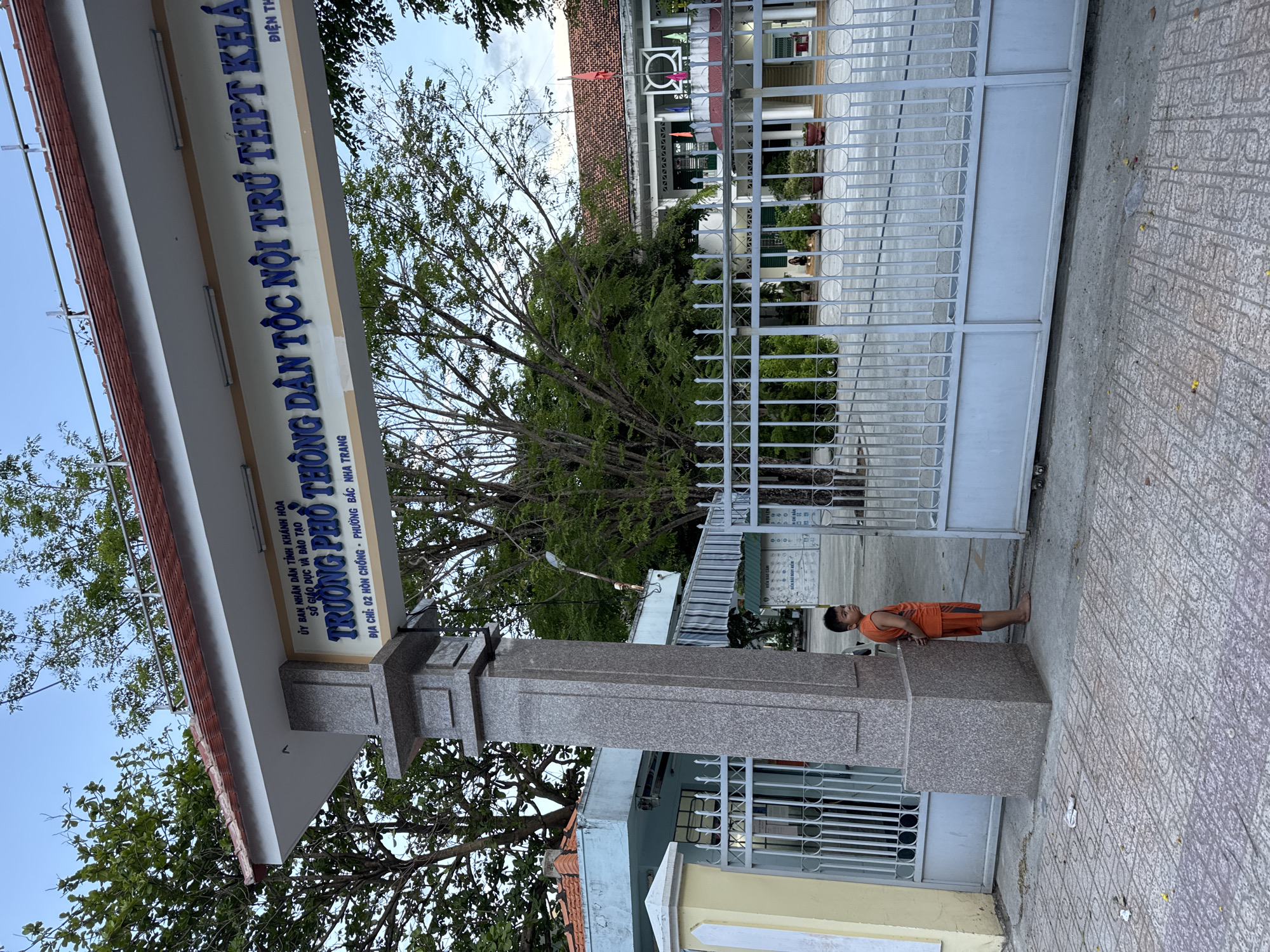 The image depicts the entrance of Trường Phổ Thông Dân Tộc Nội Trú THPT, a specialized boarding high school in Nha Trang, Vietnam, located at 02 Hon Chong, Bac Nha Trang Ward. The school sign is visible, displaying the full name and address. A child wearing an orange top and shorts stands near the entrance gate, seemingly engaging in push-ups against the gate pillar. The gate itself is made of grey metal bars. Behind the gate, the school building can be partially seen, along with some trees and foliage. The sidewalk in front of the entrance is paved with patterned tiles. The sky is visible in the upper part of the image.
