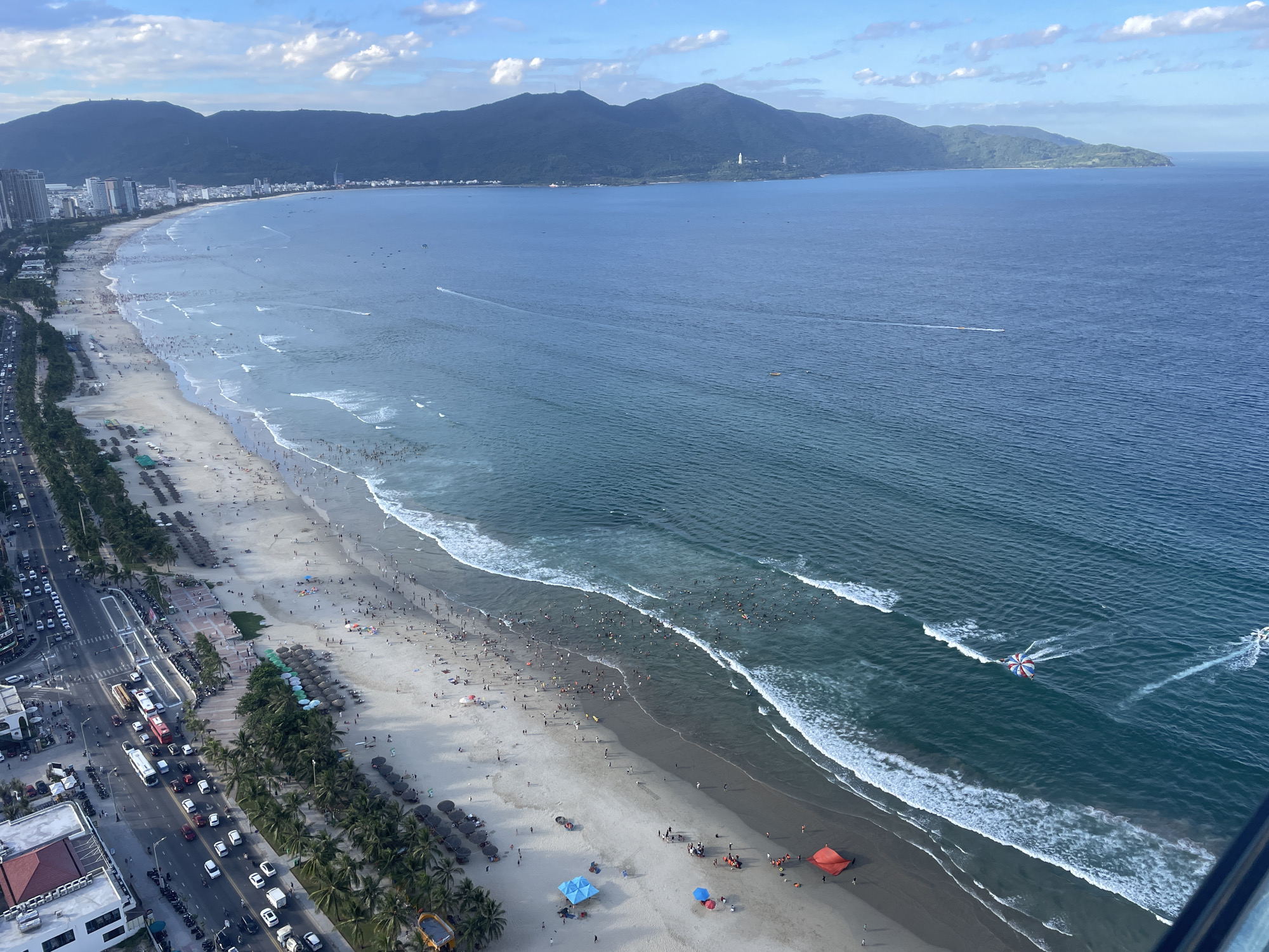 This is an aerial view of My Khe Beach in Đà Nẵng, Vietnam. A long, wide sandy beach is packed with numerous people sunbathing, swimming in the ocean, and walking along the shore. The water is a deep blue-green, with white waves breaking along the coastline. Several small boats and watercraft, including a parasail, are visible on the water.
Adjacent to the beach is a palm-lined promenade, followed by a busy multi-lane road lined with traffic, including cars, buses, and motorbikes. Tall buildings and a city skyline are visible in the distance along the coast. Behind the city, lush green mountains rise, with a cloudy blue sky overhead. The scene depicts a popular tourist destination with a vibrant atmosphere.