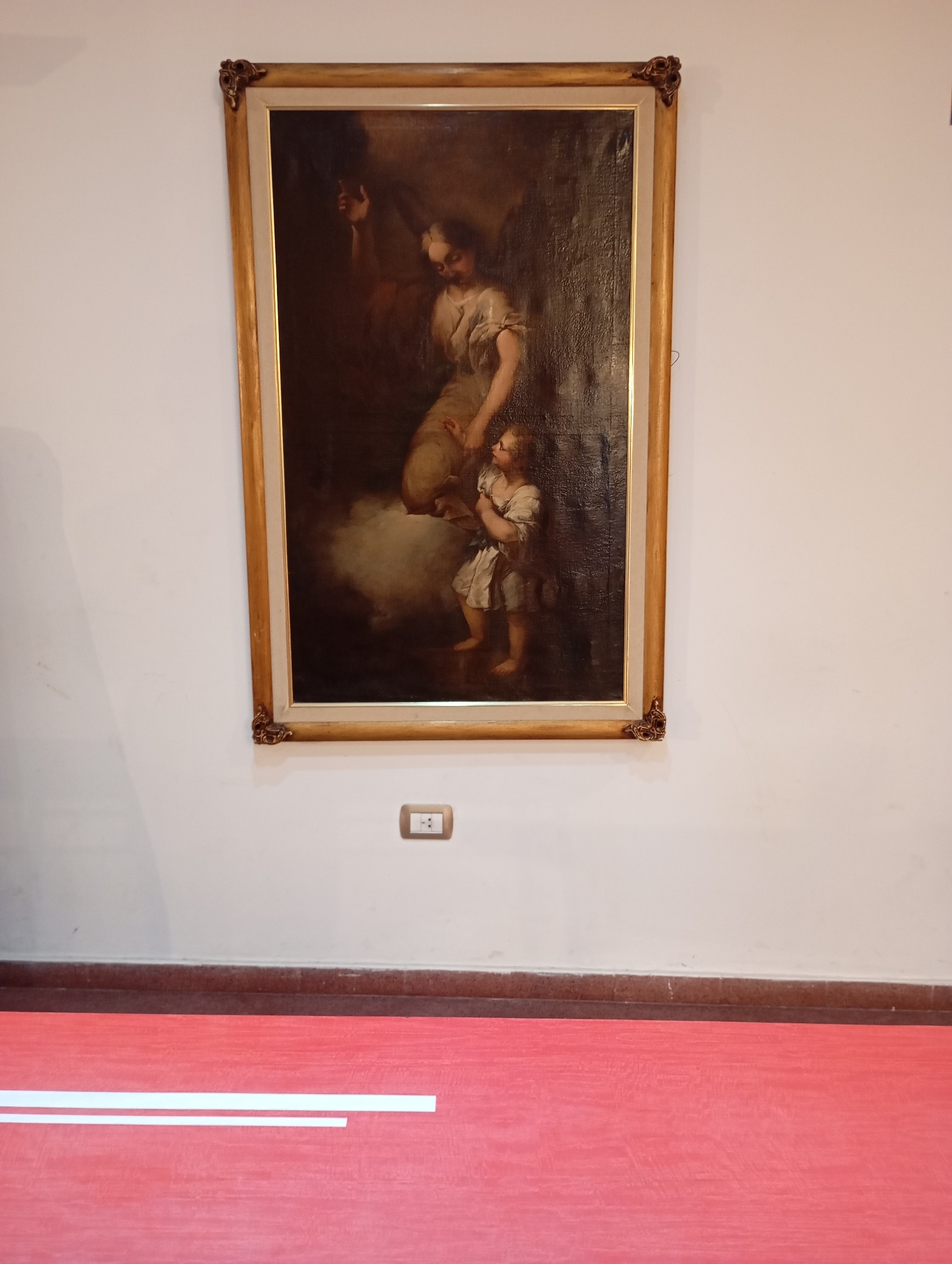The image shows a framed painting of an angel with a child. The painting is displayed on a white wall above a light-colored electrical outlet. Below the wall is a dark brown trim and then a vibrant red floor. Two white strips of material are laid parallel on the red floor. The painting is in a gold frame with ornate corners. The angel appears to be reaching out to the child, who is looking up at the angel. The scene is dimly lit, with the figures of the angel and child illuminated by a soft light. The overall impression is one of tenderness and protection. The architectural elements, such as the electrical outlet and trim, suggest an indoor setting, possibly a museum or a home in La Plata, Argentina.