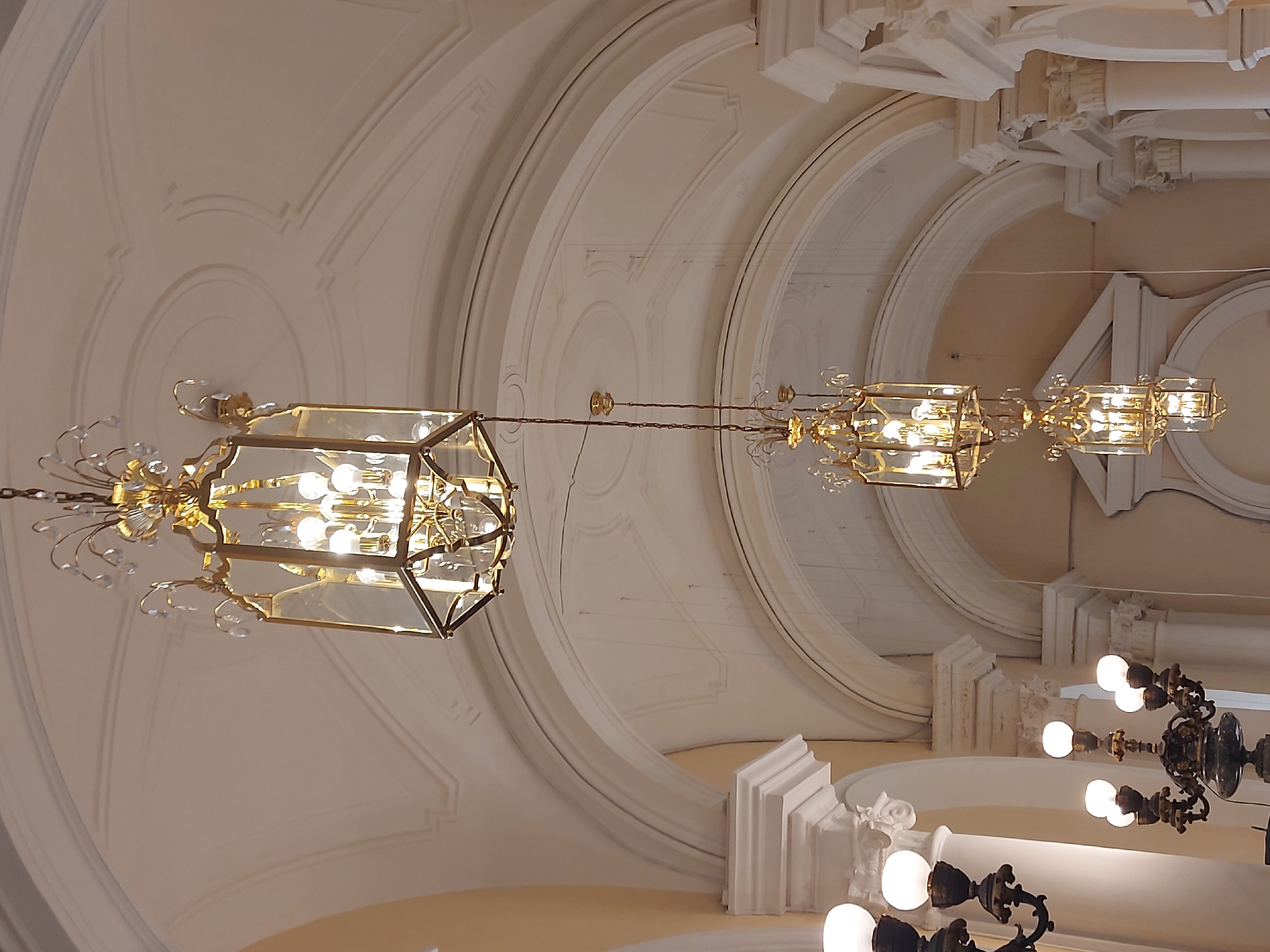 The image shows the interior of a building, potentially a palace or a grand hall, with elaborate architectural details. The ceiling is arched and adorned with ornate moldings and concentric circles. Three chandeliers of varying sizes are suspended from the ceiling. The largest, most prominent chandelier is located on the left side of the image, featuring a geometric cage design with multiple light bulbs inside. Two smaller chandeliers hang further back and to the right. Other architectural features include decorative columns and ornate detailing at the base of the ceiling. Another chandelier is partially visible in the foreground.
The photograph was likely taken at the Catherine Palace in Saint Petersburg, Russia.