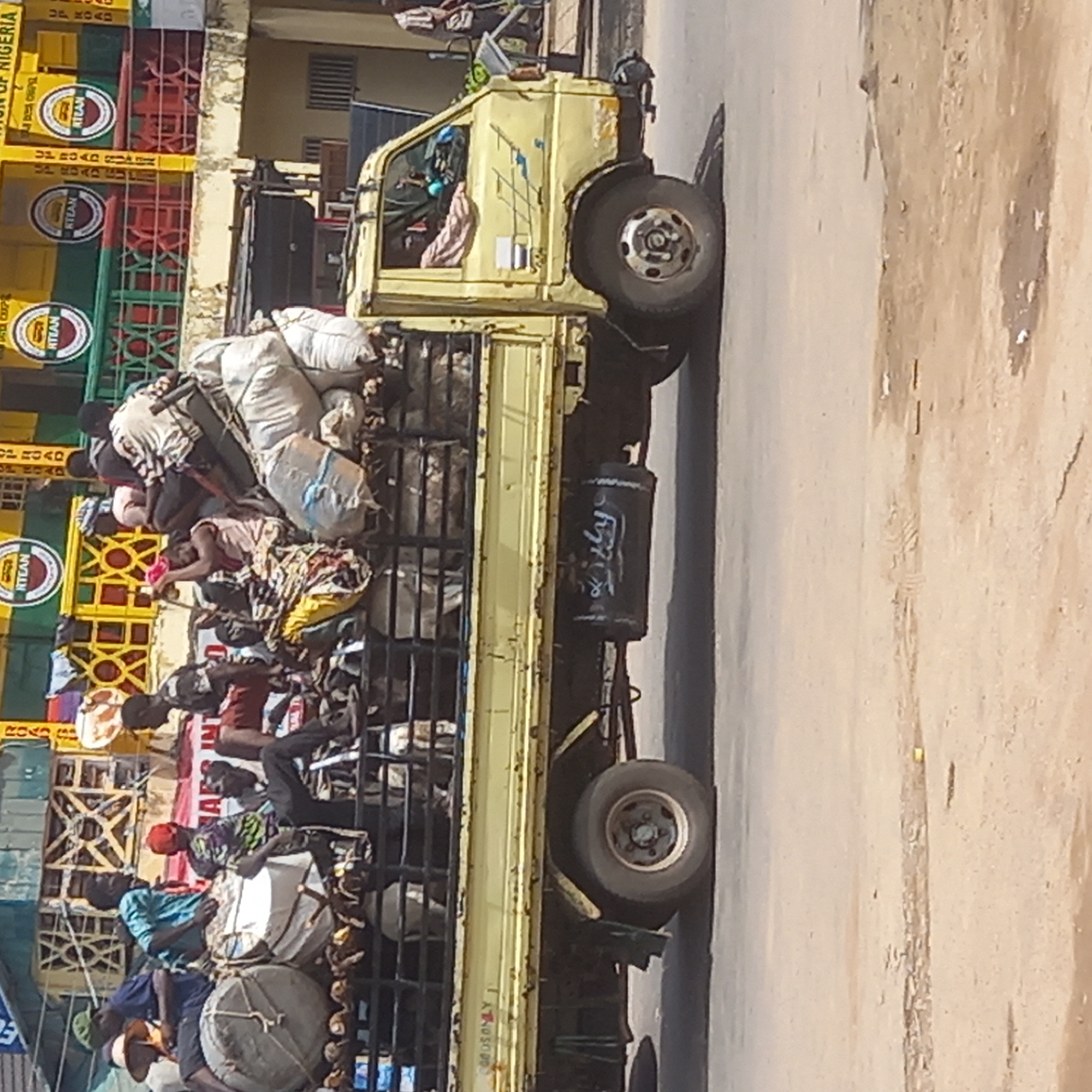 A light yellow flatbed truck travels on a paved road in what appears to be an urban area of Dadi Riba, Nigeria. The truck is heavily laden with people, sacks, and drums, all crammed into the truck bed, enclosed by metal fencing. The truck also has a cylinder, possibly an air tank, with writing on it. A building is visible in the background with signs or advertisements containing the word "RTEAN" and "NIGERIA." The scene suggests a mode of public transport or resource-sharing typical in densely populated areas.