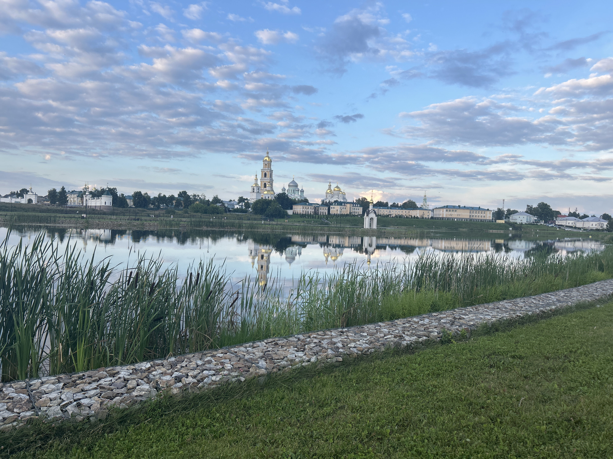 The image depicts a wide, calm body of water reflecting the sky and a distant embankment lined with buildings. The embankment features several domed structures, most notably a tall bell tower with golden domes, and other white and light-colored buildings with tiled roofs. A few green trees are scattered among the buildings. The water is bordered by a dense growth of tall green reeds and cattails along the near bank. A gabion wall made of stones fills the space between the reeds and the grassy foreground. The sky is a bright blue with scattered white, puffy clouds. The scene appears to be a serene landscape, likely a monastery or religious complex on the opposite side of the water. This location is identified as the Seraphim-Diveyevo Monastery in Diveyevo, Russia. There are no people or animals visible in the image.