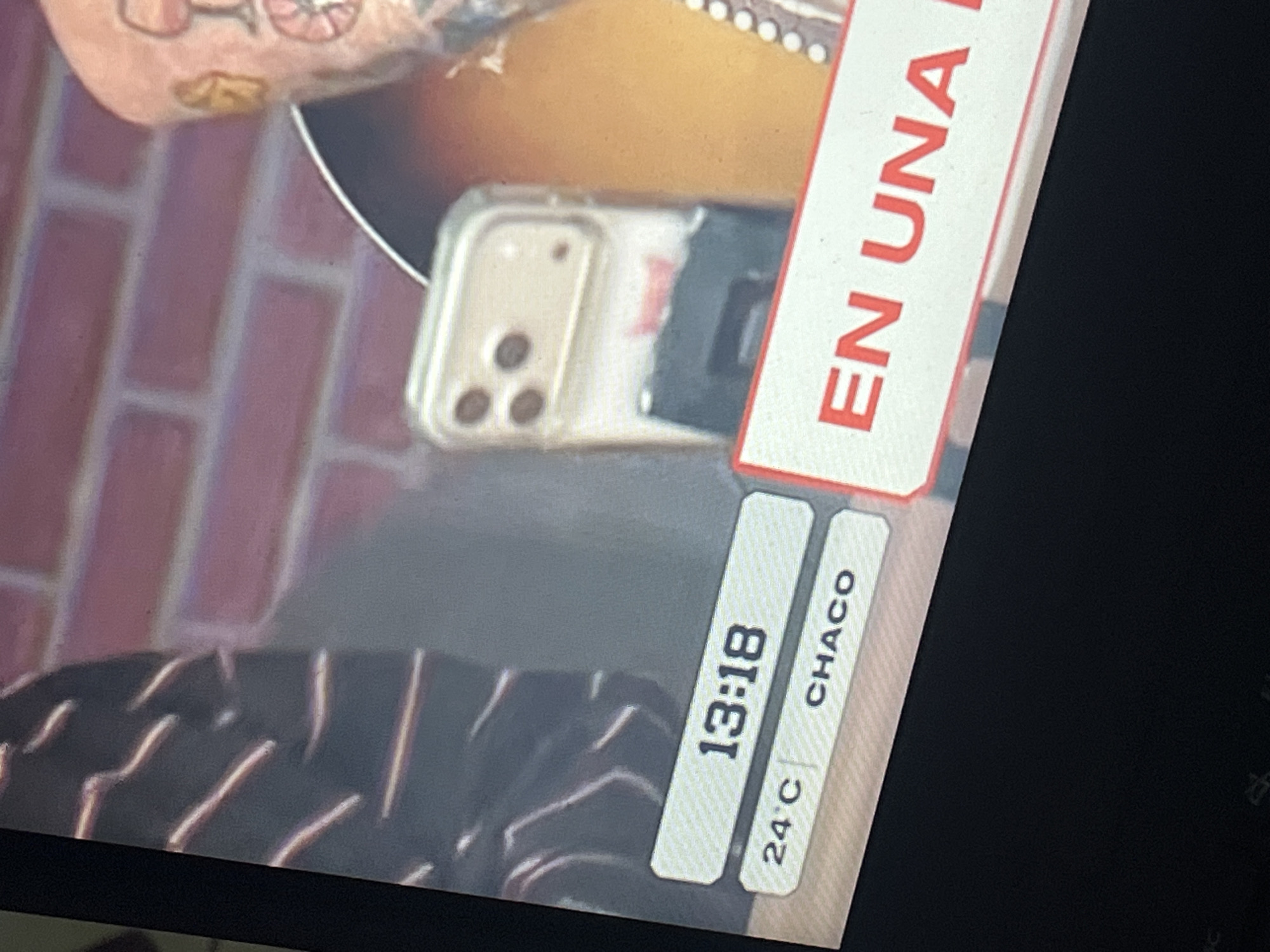 This image is a photograph of a television screen, taken in Santa Ana, Venezuela. The screen displays a broadcast featuring two individuals. On the left side of the screen, a person is visible wearing a dark garment with multiple horizontal light-colored stripes. On the right side, a second person's right arm and shoulder are visible, adorned with tattoos, including a red wine glass design and a circular spoked design beneath it. An acoustic guitar with a dark brown to orange gradient finish and white dot inlays along its edge is positioned in the lower right foreground of the broadcast. Resting on or adjacent to the guitar is a light-colored smartphone with a clear protective case, featuring a triple-lens camera array on its rear. A dark object is partially obscured beneath the phone, and a small red logo or text is blurred behind it. The background of the broadcast features a red brick wall with light-colored mortar lines. Text overlays are present on the lower portion of the screen: a white rectangular bar displaying "13:18" for the time, and below it, another white bar split into two sections showing "24°C" for temperature and "CHACO" for the regional location. In the lower right, a white rectangular banner with a red border displays partial red text "EN UNA". The very edges of a dark surface are visible at the bottom of the overall photograph, indicating the physical environment outside the TV screen.
