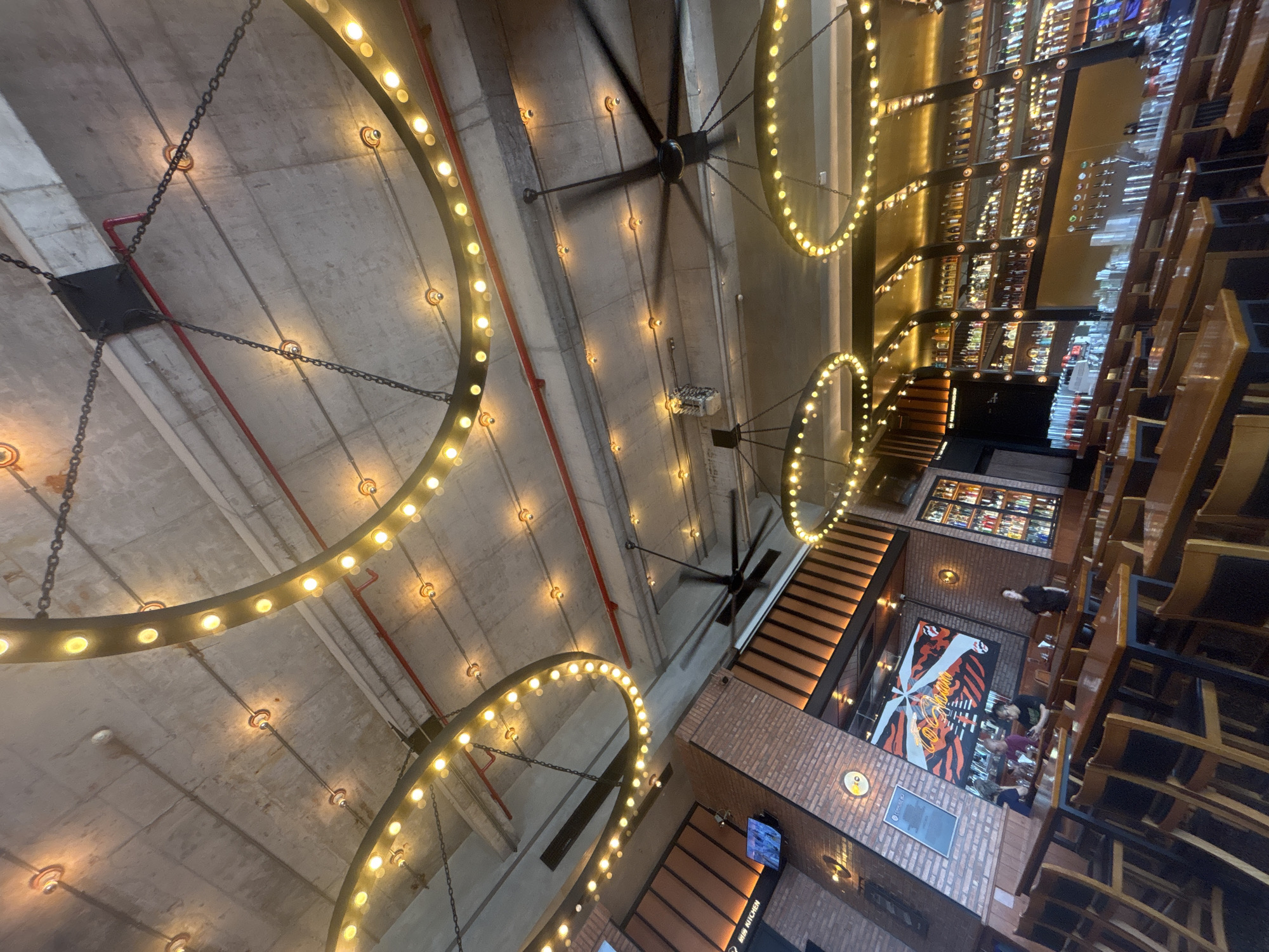 The image depicts the interior of a restaurant or bar in Nanhai District, China. The ceiling is a raw concrete slab with exposed conduits and red piping. Large, circular chandeliers adorned with numerous Edison bulbs are suspended from the ceiling by chains. A large industrial ceiling fan is also visible, spinning. The walls are clad in brick and wood paneling. Tables and chairs are arranged in the dining area, with some patrons seated at tables. A bar area is visible in the background, lined with shelves stocked with bottles of alcohol and illuminated by warm lighting. A large mural with abstract designs is mounted on one of the brick walls.