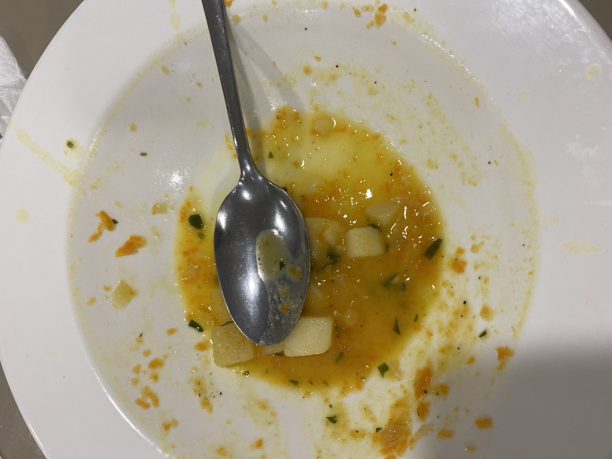 The image shows a close-up, overhead view of a white, circular plate with the remnants of a meal. A silver spoon rests in the center of the plate, partially submerged in a light orange liquid that coats the bottom of the plate. Scattered throughout the plate are small, diced pieces of what appear to be potato or similar root vegetable, as well as flecks of orange puree and green herbs, likely from a soup or stew. The edges of the plate are stained with the same orange residue. The background is out of focus, but appears to be a tabletop with a white napkin to the upper right. There are no people or animals visible in the image. The scene depicts the aftermath of a meal, with only residue remaining.