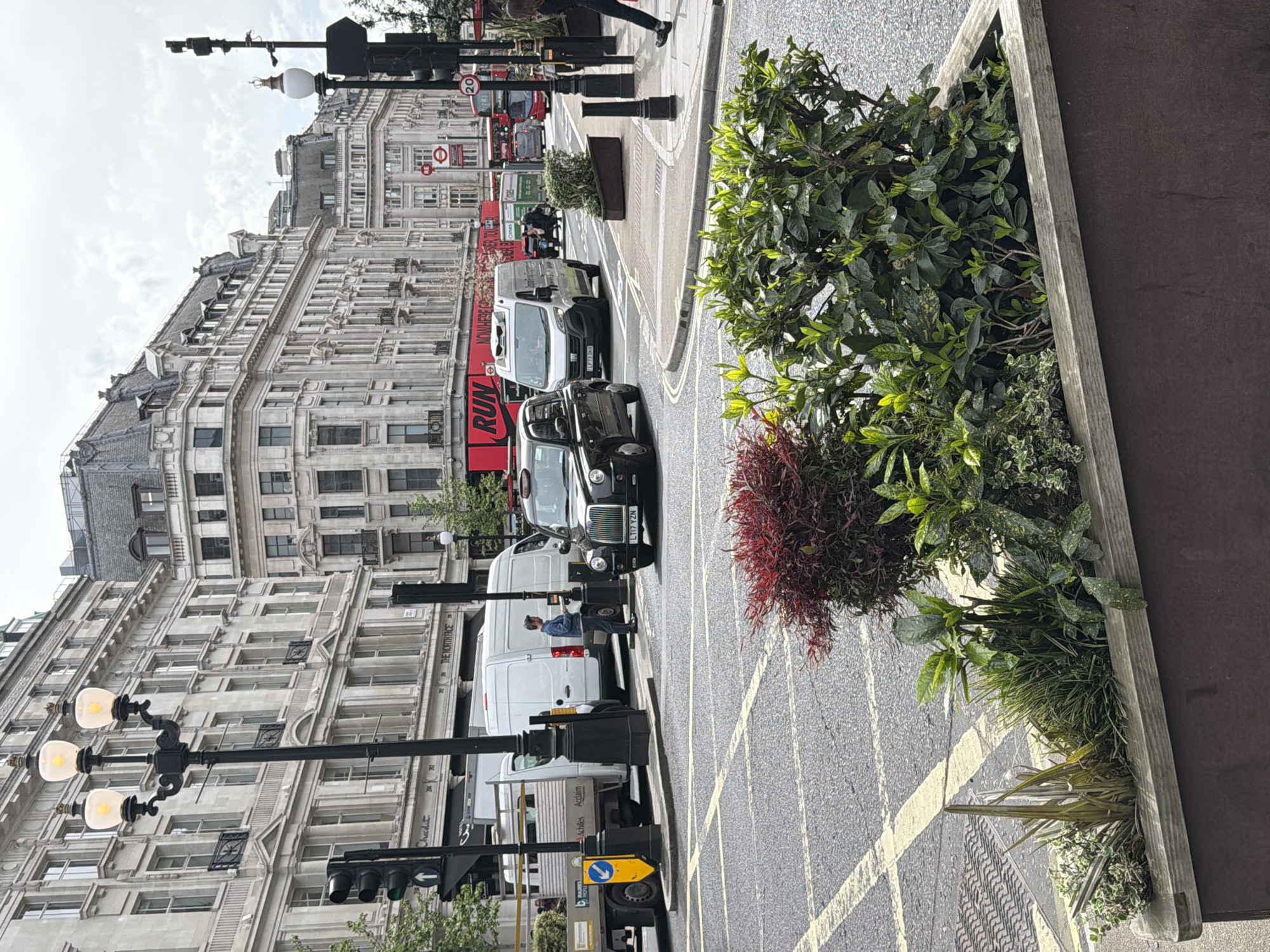 The image depicts a street in London, United Kingdom. Buildings with multiple windows line the street on the left, and traffic including black cabs and vans fill the road. Pedestrians can be seen on the sidewalk. In the foreground, a raised planter box filled with greenery and red foliage is positioned next to the sidewalk. Yellow lines are painted on the street, and street lights and traffic signals are present.