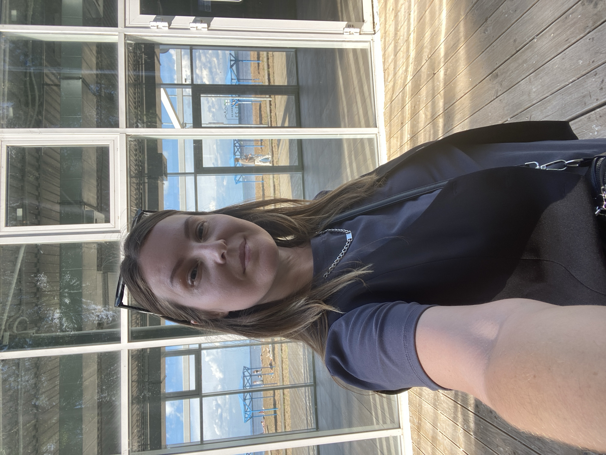 A woman takes a selfie in an outdoor setting. She is positioned in the lower left of the frame, facing the camera. She has long, light brown hair and is wearing glasses, a dark top, and a dark vest. A silver chain necklace is visible. The background is dominated by large glass windows reflecting the sky and a wooden deck. Through the windows, a body of water, a beach area, and a blue structure with what appear to be animals or people are visible. The lighting suggests it is daytime, possibly late afternoon due to the long shadows on the wooden deck. The overall mood appears casual and relaxed. The location context is Chaikovsky, Russia.