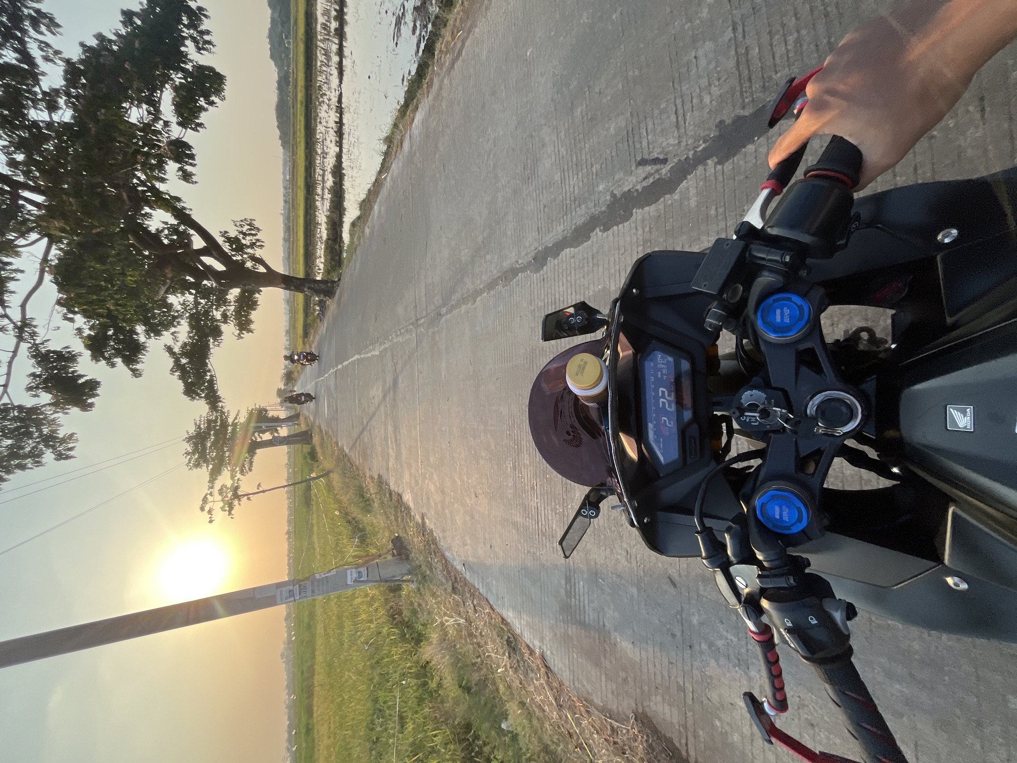 A first-person perspective shot from a motorcycle in Pecangaan, Indonesia, shows a paved road with a field of rice paddies on the right and a green field on the left. The sun is setting, casting a warm glow over the landscape. A utility pole stands to the left of the road, and trees line the right side. Several motorcycles are visible in the distance, traveling along the road. The motorcycle in the foreground has a digital display showing "22 22". A person's hand is visible on the right handlebar. The scene depicts a typical rural Indonesian setting during golden hour.