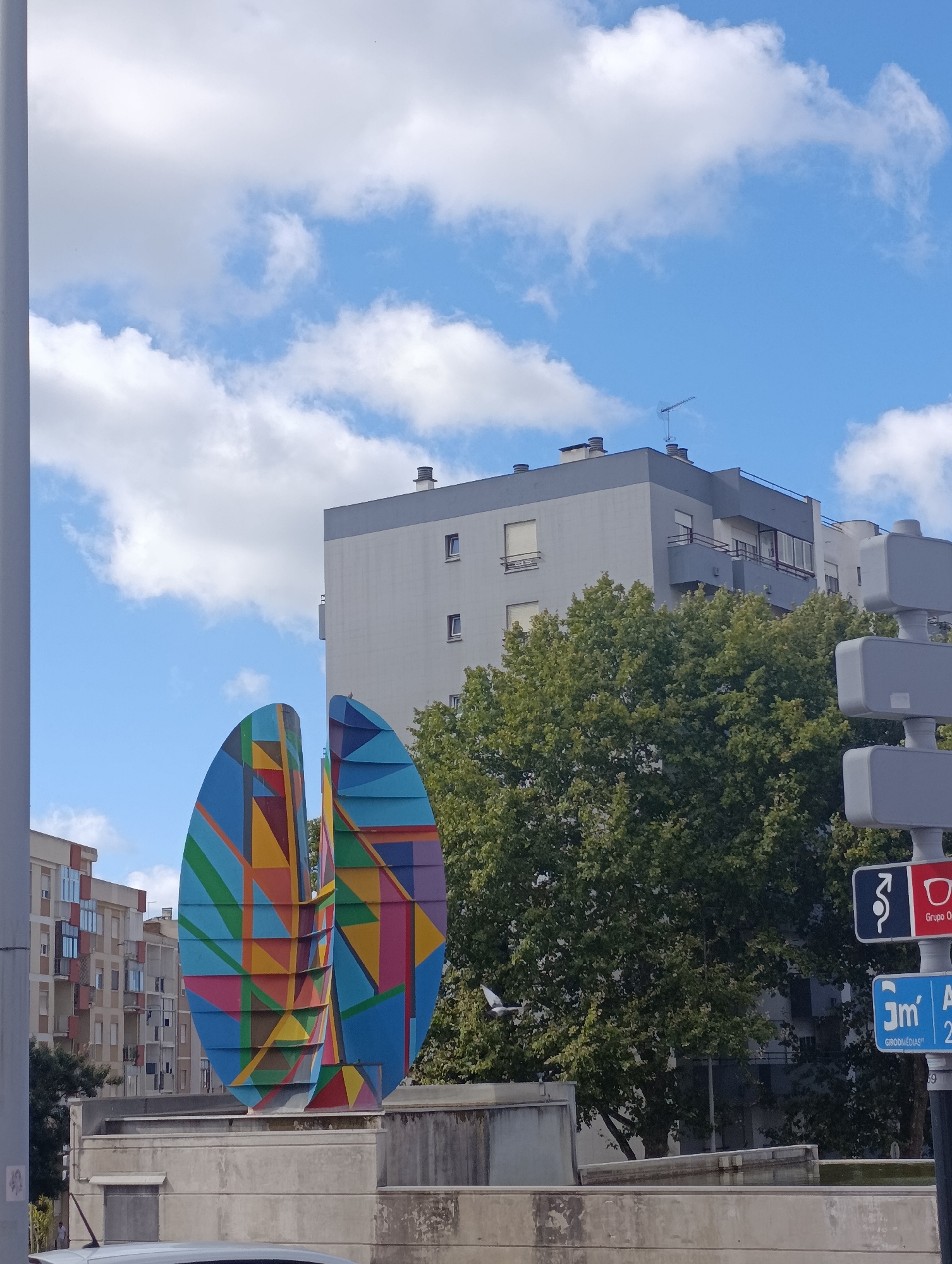 A brightly colored, geometric sculpture stands in front of a modern apartment building under a bright blue sky with scattered clouds. The sculpture, composed of interlocked, triangular shapes in vibrant blues, yellows, reds, and greens, is positioned on a raised concrete platform. Behind it, a medium-height apartment building with gray and white sections and balconies is visible. To the right of the sculpture, lush green trees partially obscure the view of the building. A street sign with multiple rectangular panels, some bearing red and blue backgrounds with white symbols and text, is mounted on a pole to the right of the frame. The lower portion of the image shows a concrete barrier and a sliver of a white car. The scene appears to be an urban environment in Setúbal, Portugal, possibly a public square or street.