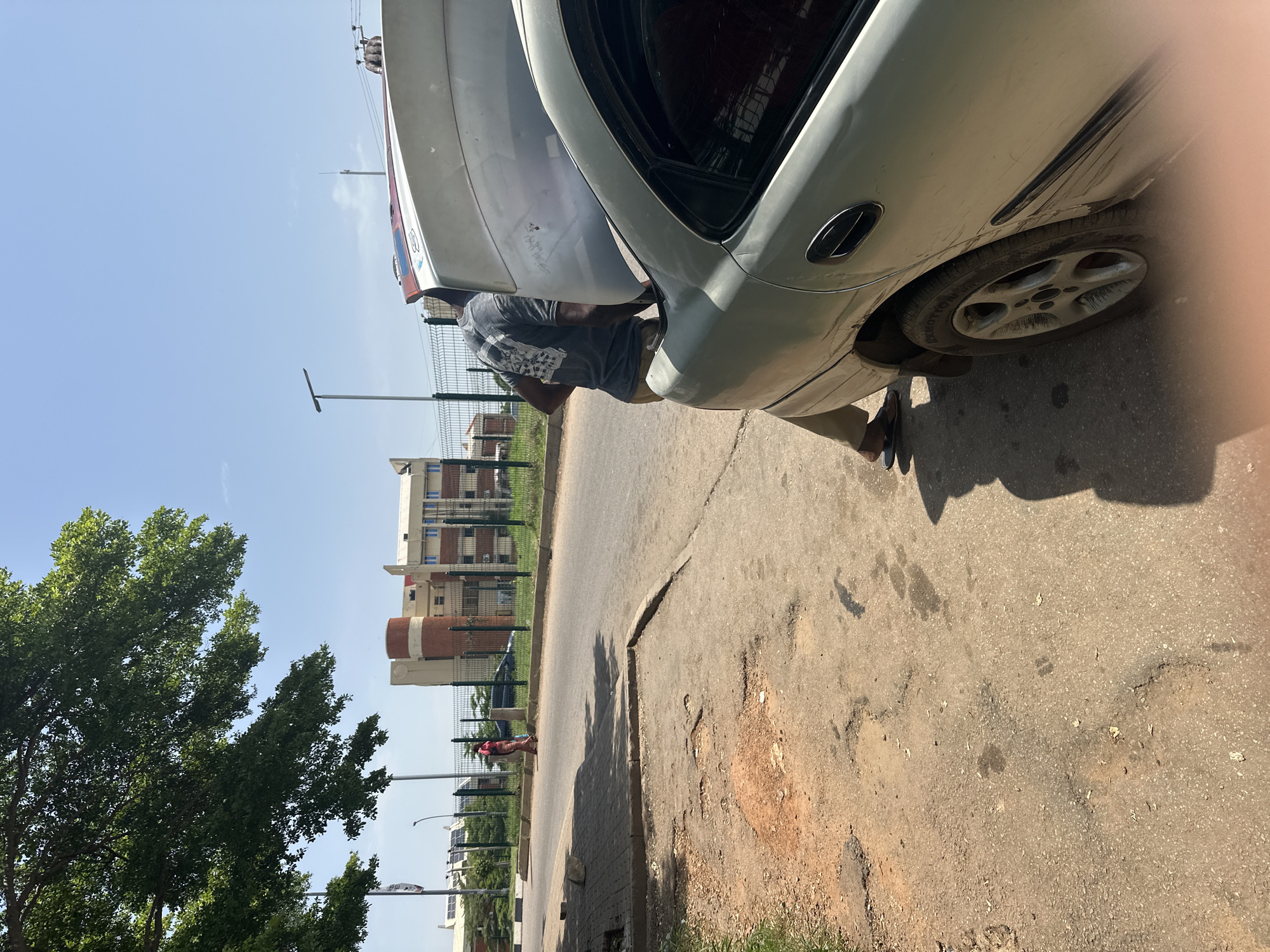 The image depicts an outdoor scene on a sunny day in Abuja, Nigeria. A man, wearing a dark t-shirt and light-colored pants, is seen working on the rear of a light-colored car. The car appears to be a sedan, and the trunk is open, with a large white object, possibly a spare tire or another car part, resting on top of it. The man is positioned behind the car, appearing to be engaged in some form of repair or maintenance.
The setting is a roadside with cracked asphalt pavement and a patch of dry, reddish earth to the right. A tall, leafy green tree stands to the left of the frame. In the background, there are several multi-story buildings, characteristic of an urban environment, and a green metal fence runs along the side of the road. Streetlights and power poles are also visible. The sky is clear and blue, indicating fair weather. The overall impression is a common scene of roadside vehicle service.