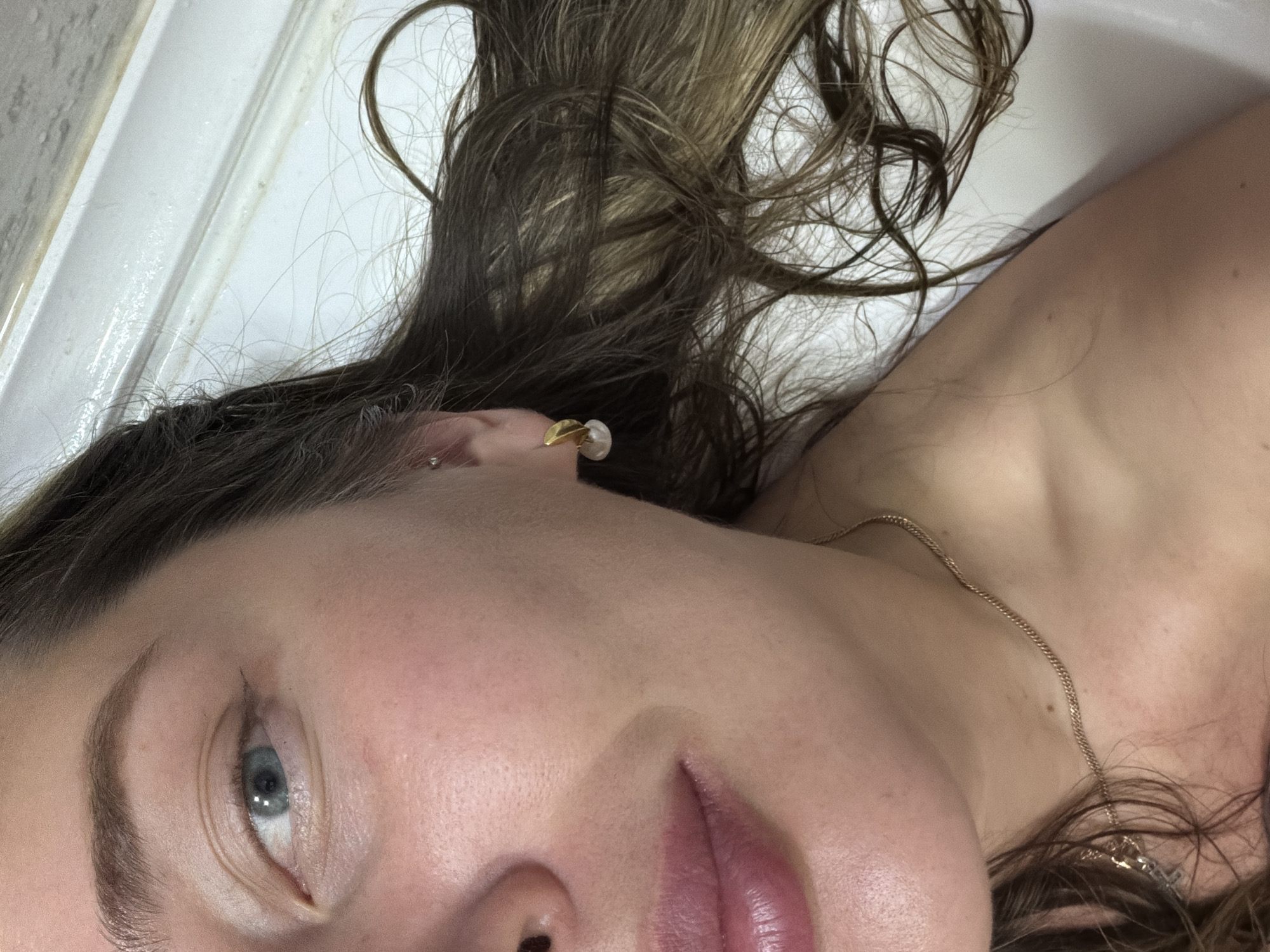 A close-up, tilted image of a woman's face, appearing to be lying down, possibly in a bathtub. Her hair is a mix of brown and blonde, styled with loose curls and some strands framing her face. She is wearing an earring, which looks like a gold stud with a white pearl. A thin gold chain necklace is visible around her neck. The background includes a white, glossy surface, likely the inside of a bathtub or a similar fixture. The woman's eye is light-colored. No specific location in Chaikovsky, Russia, can be determined from the given image.