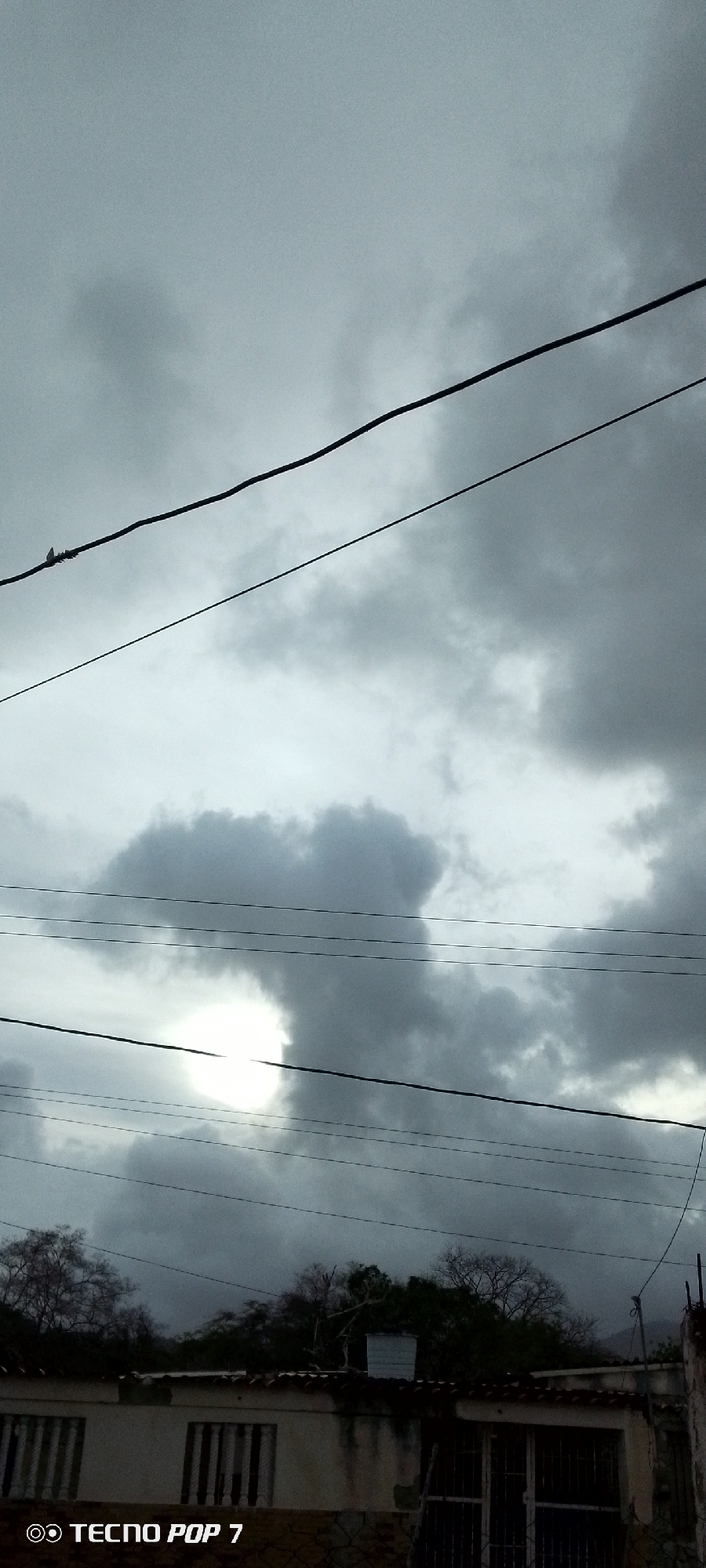 The image, taken with a TECNO POP 7 phone, depicts a cloudy sky above a building in Altagracia, Venezuela. Several electrical wires run across the frame, partially obscuring the view of the clouds. The building is a single-story structure with barred windows. Trees are visible behind the building. The overall tone is somber due to the overcast conditions. A bird is perched on one of the power lines.
