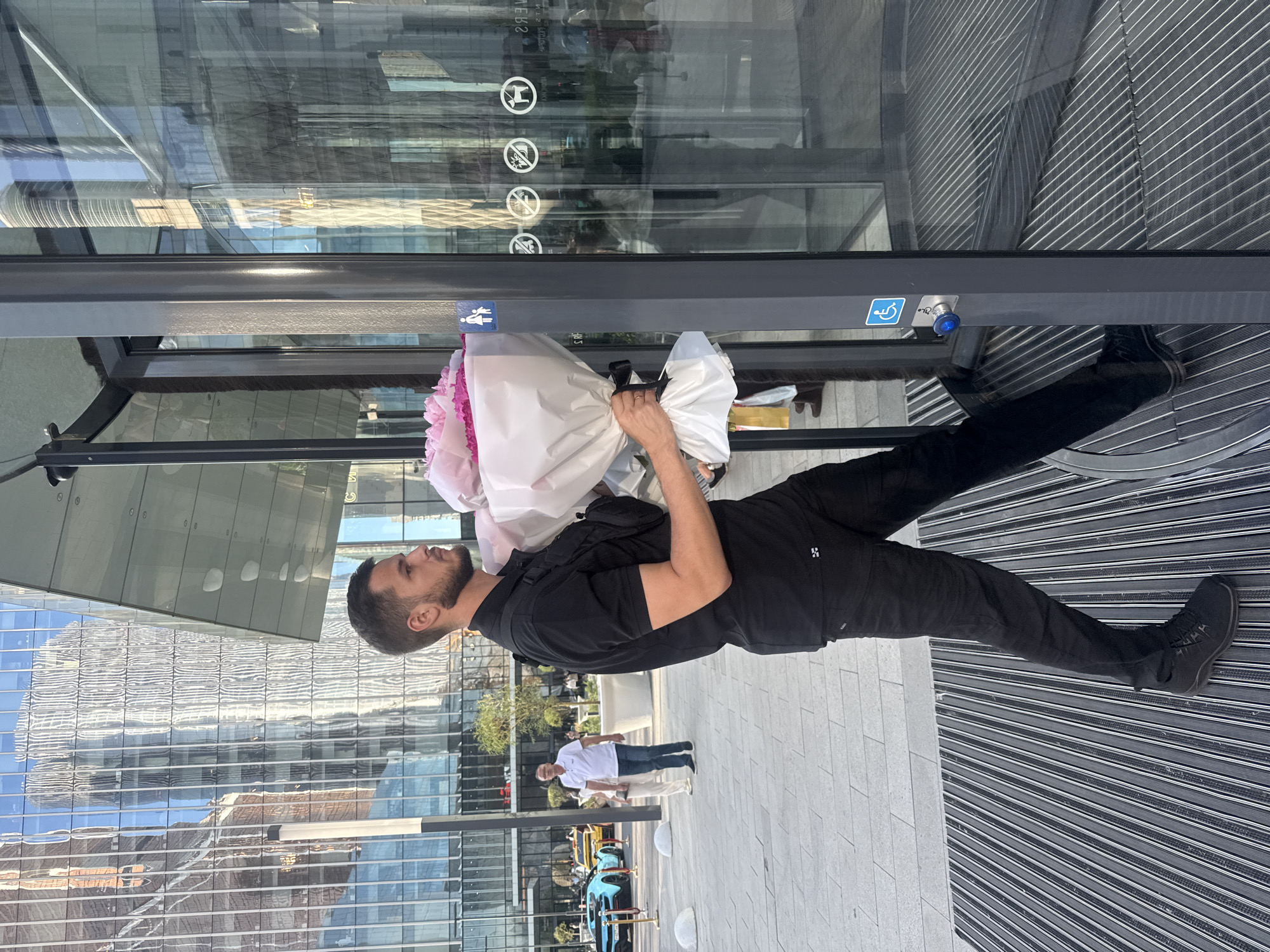 A man in black clothing is walking into a revolving glass door with a bouquet of pink and white flowers. He is wearing black pants, a black short-sleeved polo shirt, and black sneakers. He has short dark hair and a beard. He is carrying the flowers in his right hand, which is partially obscured by the bouquet. A black fanny pack is worn across his chest. The revolving door is part of a modern building with a glass facade, reflecting the surrounding cityscape. Outside the building, there are people, cars, and paved walkways. The image appears to be taken in the city of Khimki, Russia, given the context of the surrounding buildings and urban environment.