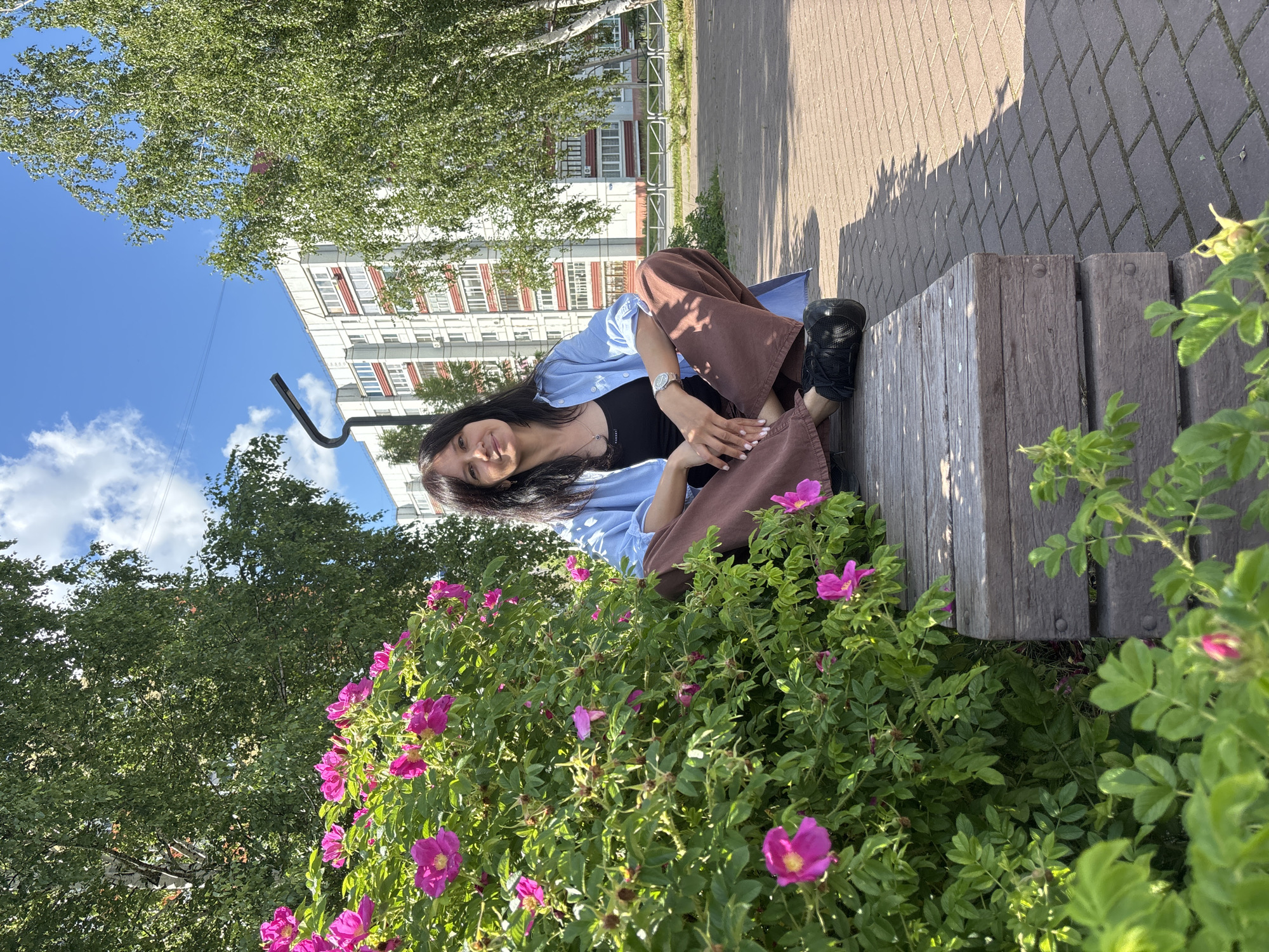 A woman with dark hair is sitting cross-legged on a wooden bench in a park. She is wearing a light blue open shirt over a black tank top and brown wide-leg pants, with black sneakers. She is smiling at the camera, with her hands resting on her lap. To her left is a large bush with bright pink flowers. In the background, a white and red apartment building stands tall, surrounded by green trees. The ground is paved with brick. The scene appears to be in a residential area of Chaikovsky, Russia, likely a public green space adjacent to apartment buildings. The sky is blue with scattered clouds, indicating a sunny day.