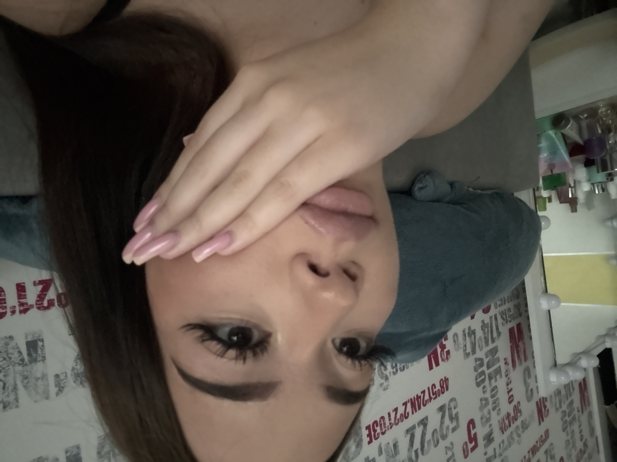 A selfie captures a fair-skinned person with brown hair, positioned at an angle. The individual's right hand partially obscures their face, revealing the eyes and mouth. Makeup is visible, particularly on the eyes. The background consists of a bed with printed text and a vanity table featuring various bottles. While the image context does not mention Naberezhnye Chelny, Russia, the printed text in the background seems to refer to coordinates (latitude and longitude), but they are insufficient to confirm a specific location in Naberezhnye Chelny, Russia.
