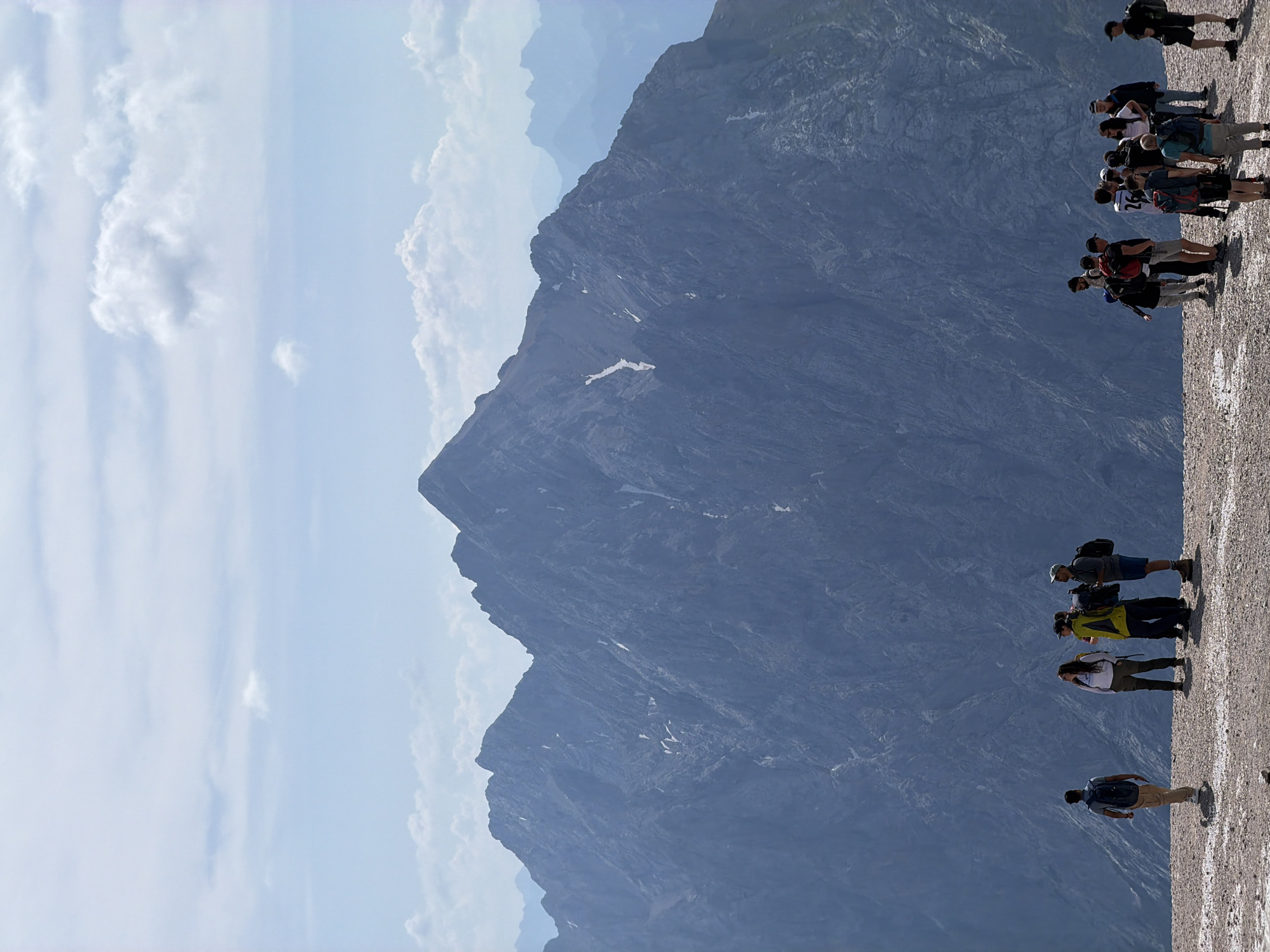 A group of hikers are gathered on a rocky, elevated area with a vast, imposing mountain range in the background. The sky is a pale blue with scattered white clouds. The hikers, varying in age and attire, are mostly facing away from the camera, some looking towards the mountains or interacting with each other. Several individuals are wearing backpacks, suggesting they are on a trek. The foreground is a textured, uneven surface of stones and pebbles. The mountains are rugged and steep, with patches of snow visible on their slopes. The scene evokes a sense of adventure and awe inspired by the natural landscape. This location is the Zugspitze massif, Germany's highest peak, which can be accessed from Garmisch-Partenkirchen.