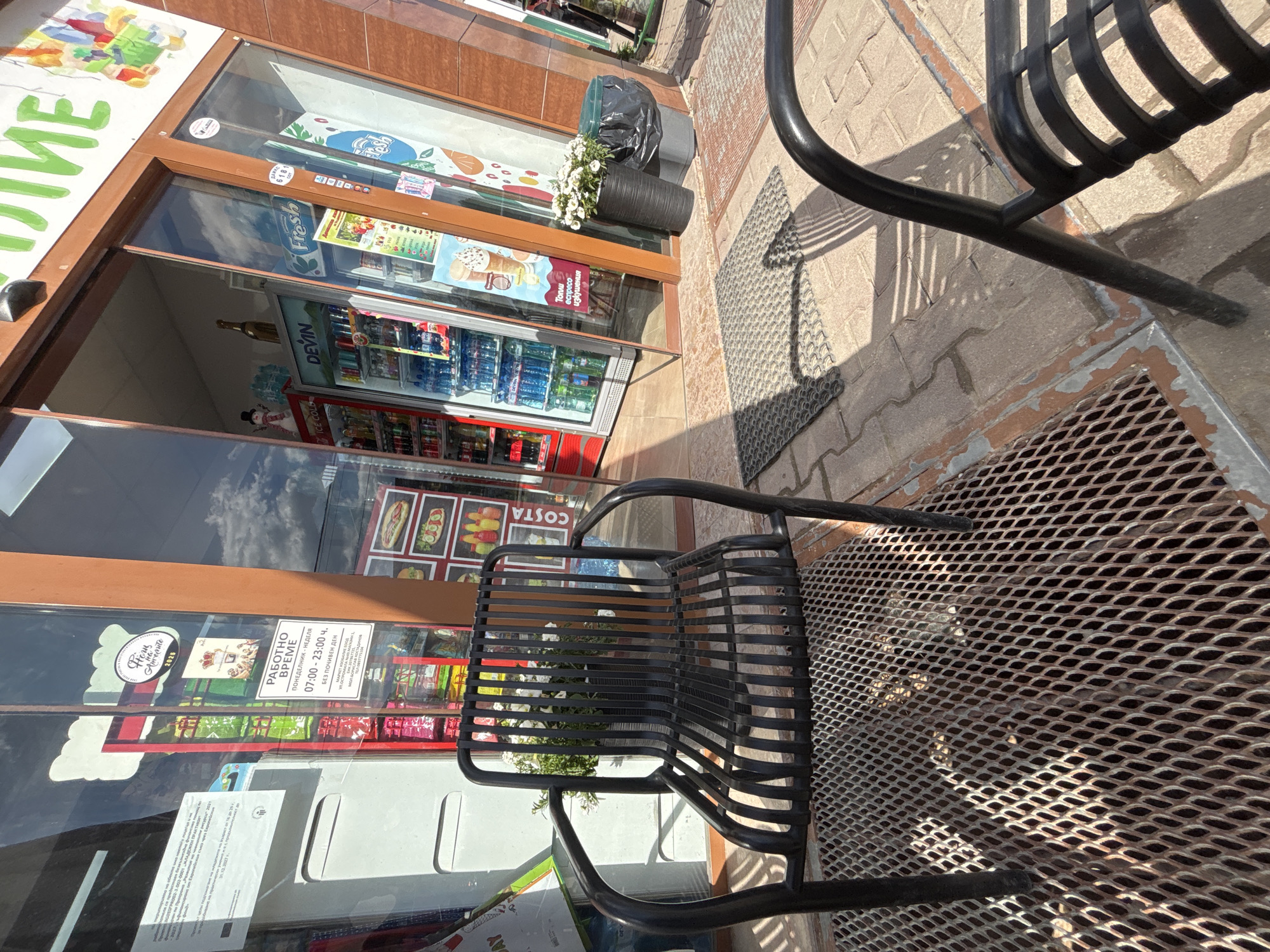 This image captures an outdoor scene at a storefront in Plovdiv, Bulgaria. A black metal chair sits on a patterned metal grate. The storefront features a glass window displaying various items for sale, including beverages and food products. A sign in the window indicates the business hours as 07:00-23:00, Monday to Sunday, without a day off. A Costa sign is also visible. A flower pot is placed to the side of the store.