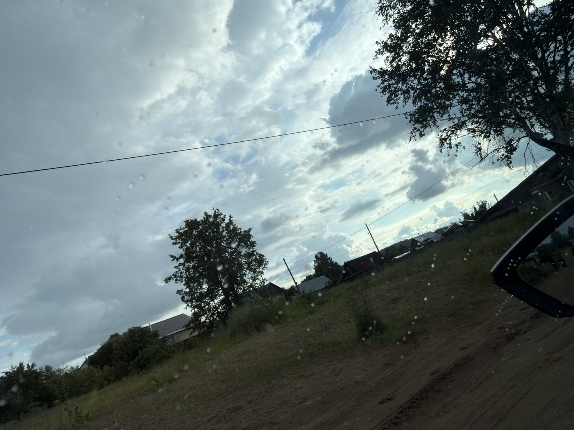 The image captures a rural landscape with a view of a residential area under a cloudy sky. The foreground consists of a dirt road with tire tracks, indicating it is a well-traveled path. To the right, the side mirror of a vehicle is visible, reflecting a bit of the scene. Beyond the road, a grassy area slopes upwards towards a cluster of houses. These houses appear to be single-story structures with dark roofs. Some trees and dense vegetation surround the houses, partially obscuring them. In the background, power lines stretch across the frame, supported by utility poles. The sky is filled with a mix of bright white and dark grey clouds, suggesting an overcast or potentially rainy day. The overall impression is of a quiet, rural setting. The image is taken from inside a vehicle, looking out through a window, as evidenced by the water droplets on the glass. The location is the city of Chaikovsky, Russia.